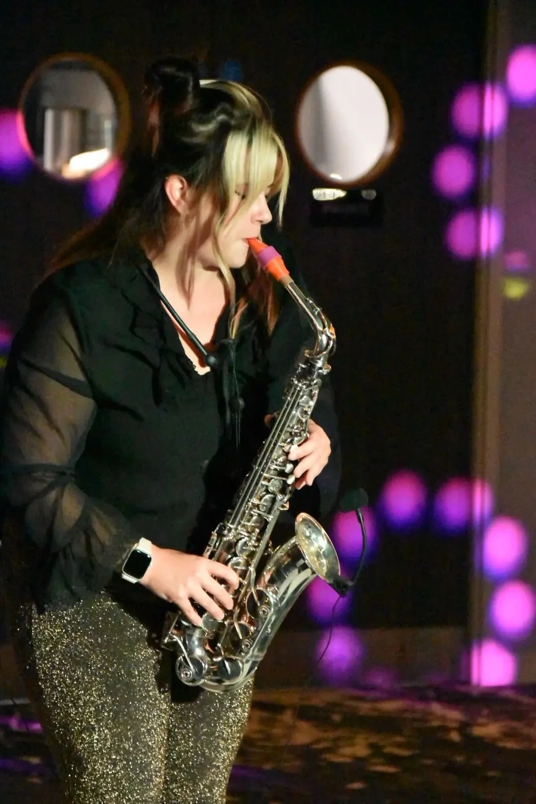 Woman playing a silver saxophone on a dimly lit stage with purple lights