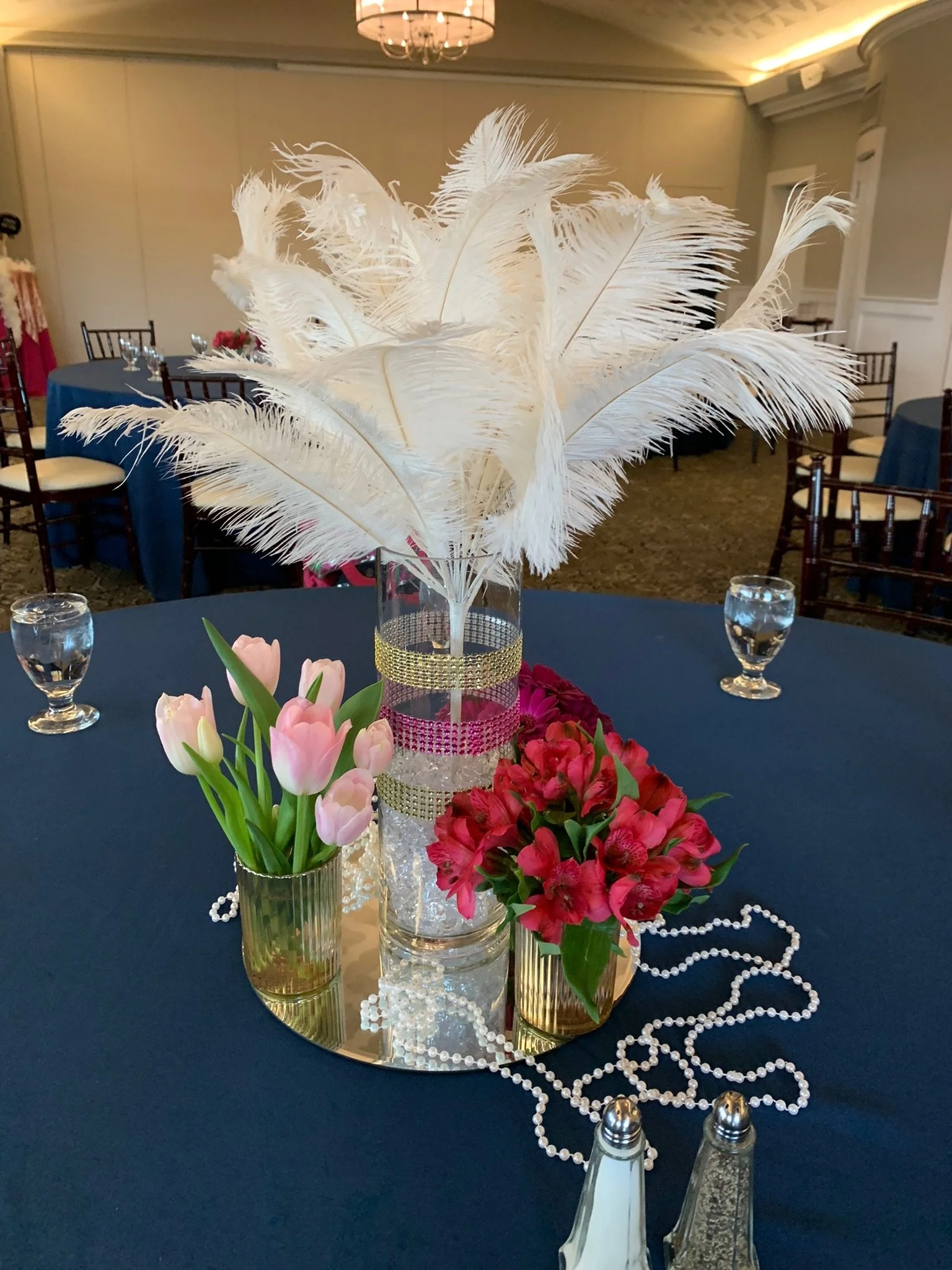 Table centerpiece with white feathers in a clear glass vase, surrounded by pink tulips, red flowers, pearl beads, and gold vases, on a dark tablecloth. Water glasses and salt and pepper shakers are also on the table, in a decorated banquet room.