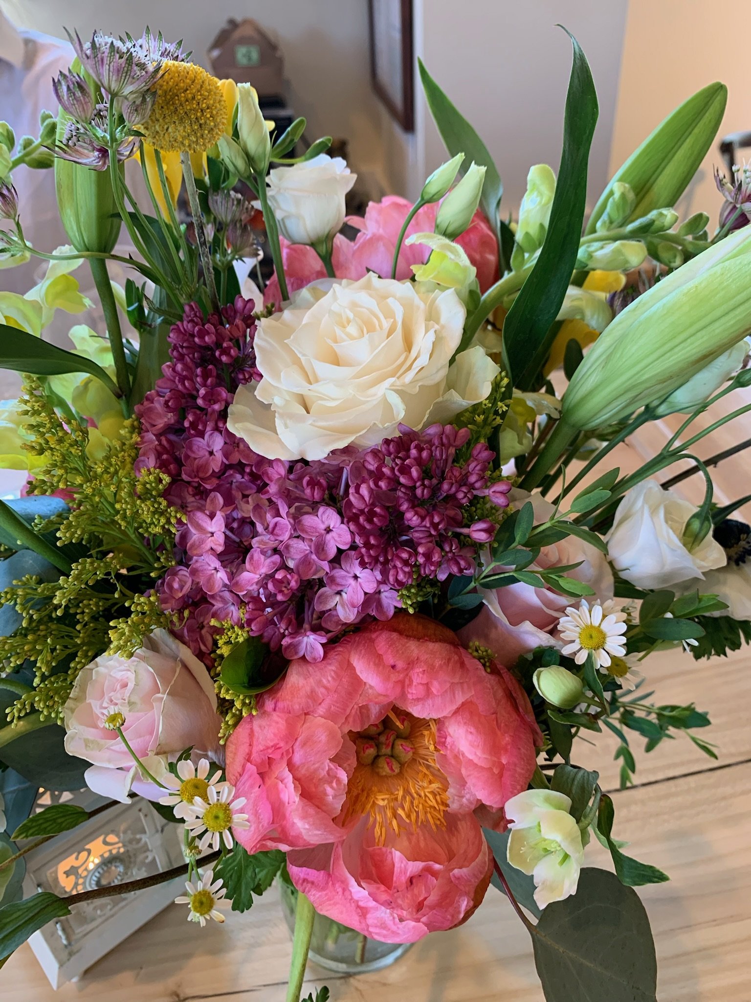 A colorful bouquet of various flowers in a glass vase, including white roses, pink peonies, purple lilacs, white daisies, and green leaves, sitting on a wooden surface indoors.