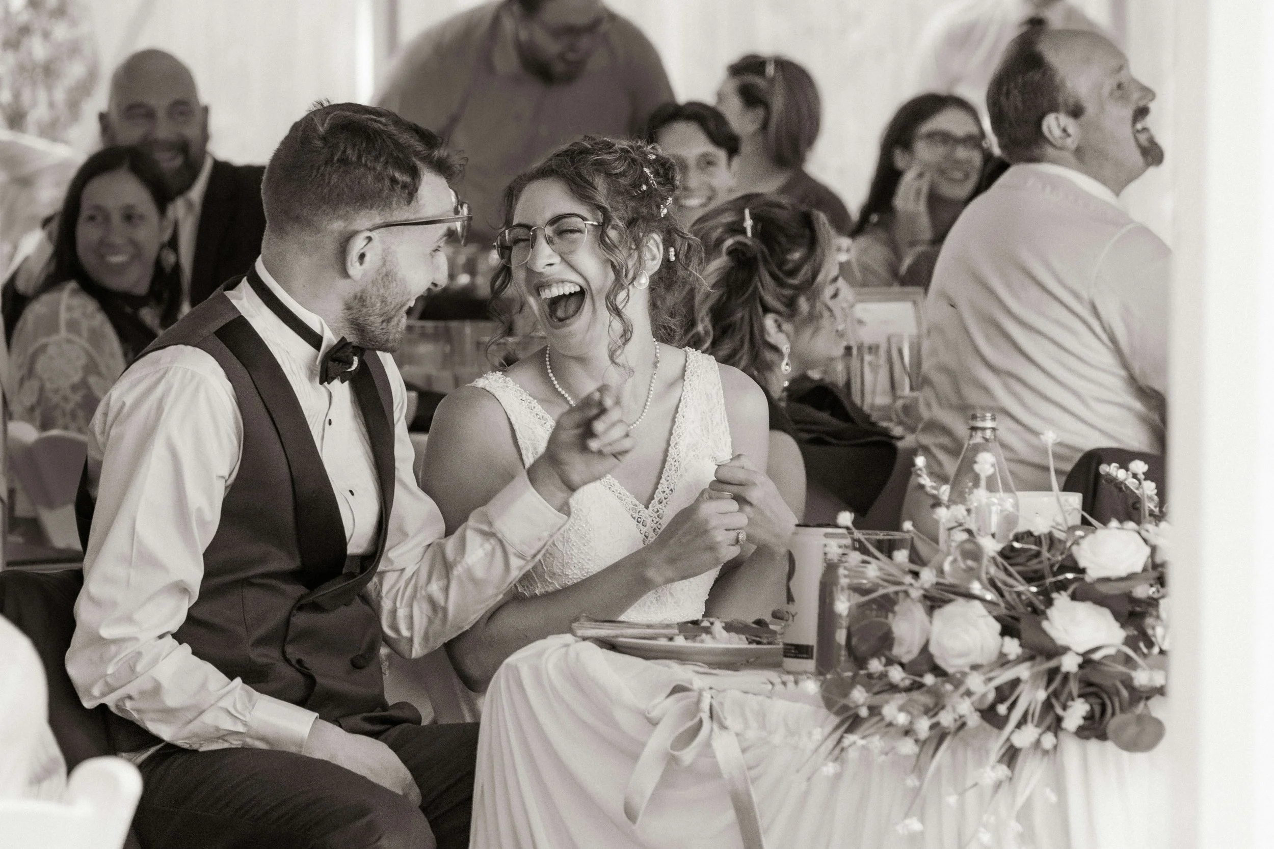A group of people at a wedding reception, laughing and enjoying himself. The bride and groom are seated at a table with flowers and drinks.