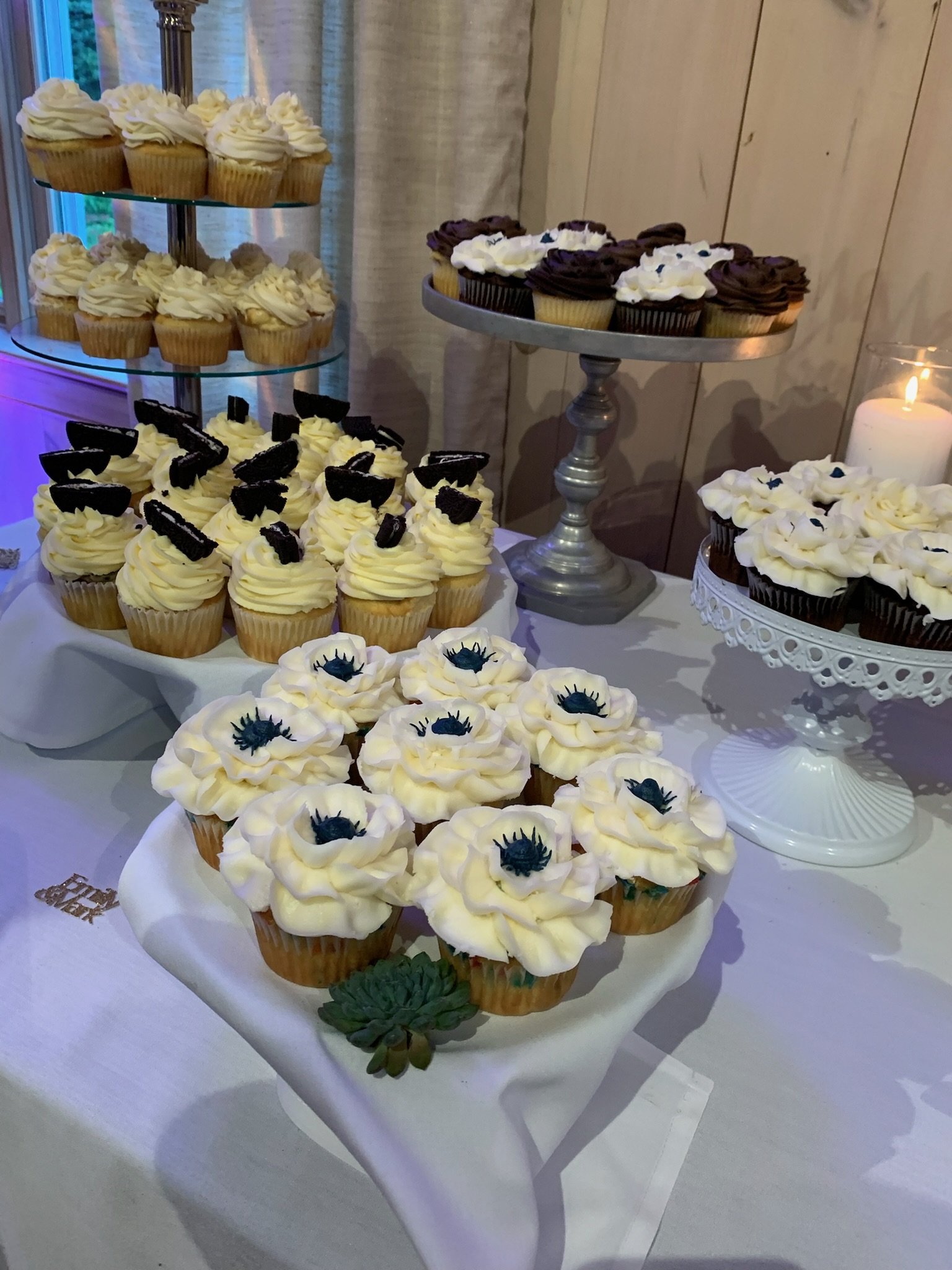 Assorted cupcakes on a dessert table, decorated with white, chocolate, and cookies and cream toppings, with some topped with black candies designed to look like spider eyes.