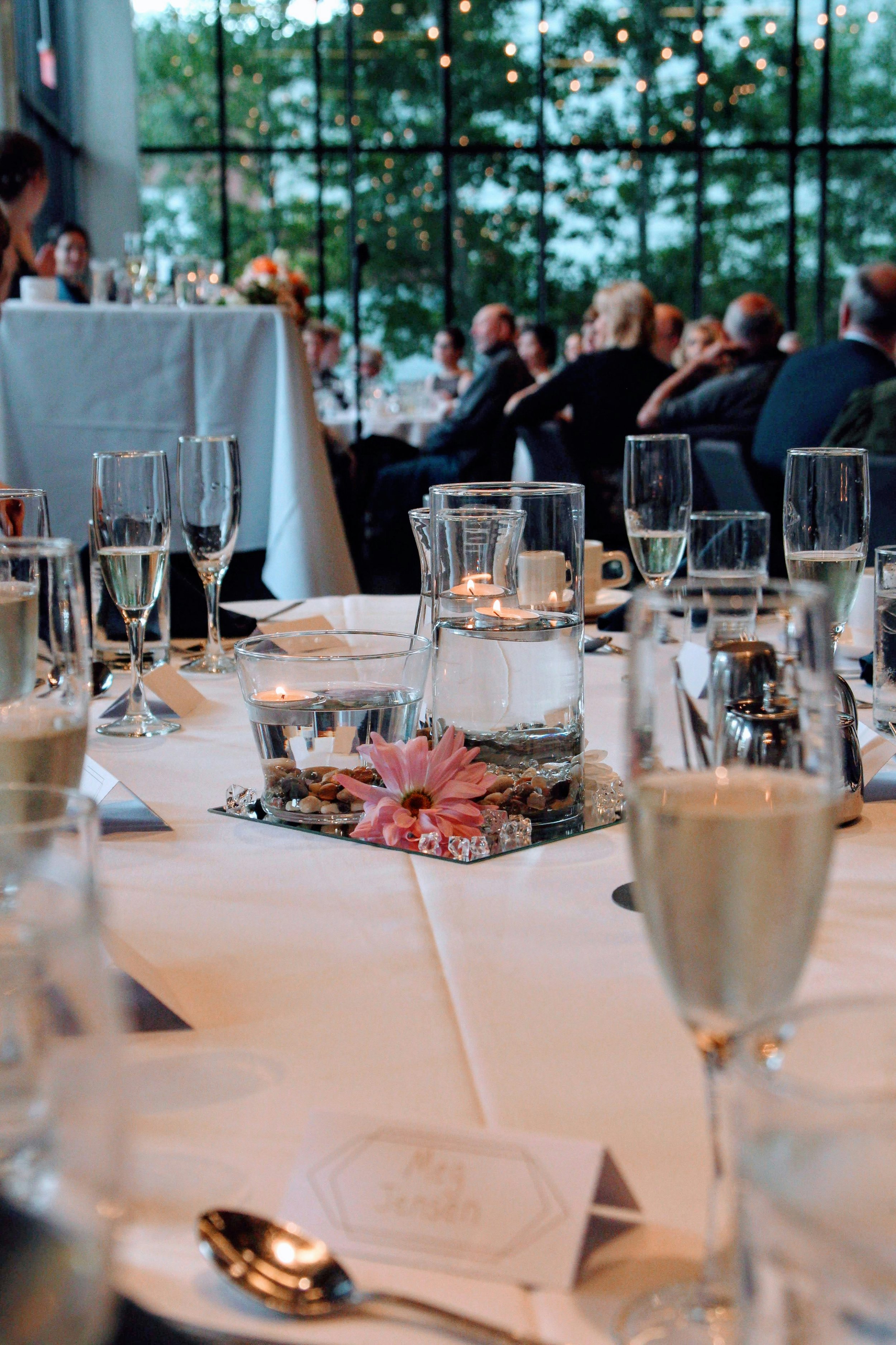 A banquet table set for a formal event with champagne flutes, a floral centerpiece, candles, and silverware. In the background, people are seated in front of a large window with string lights.