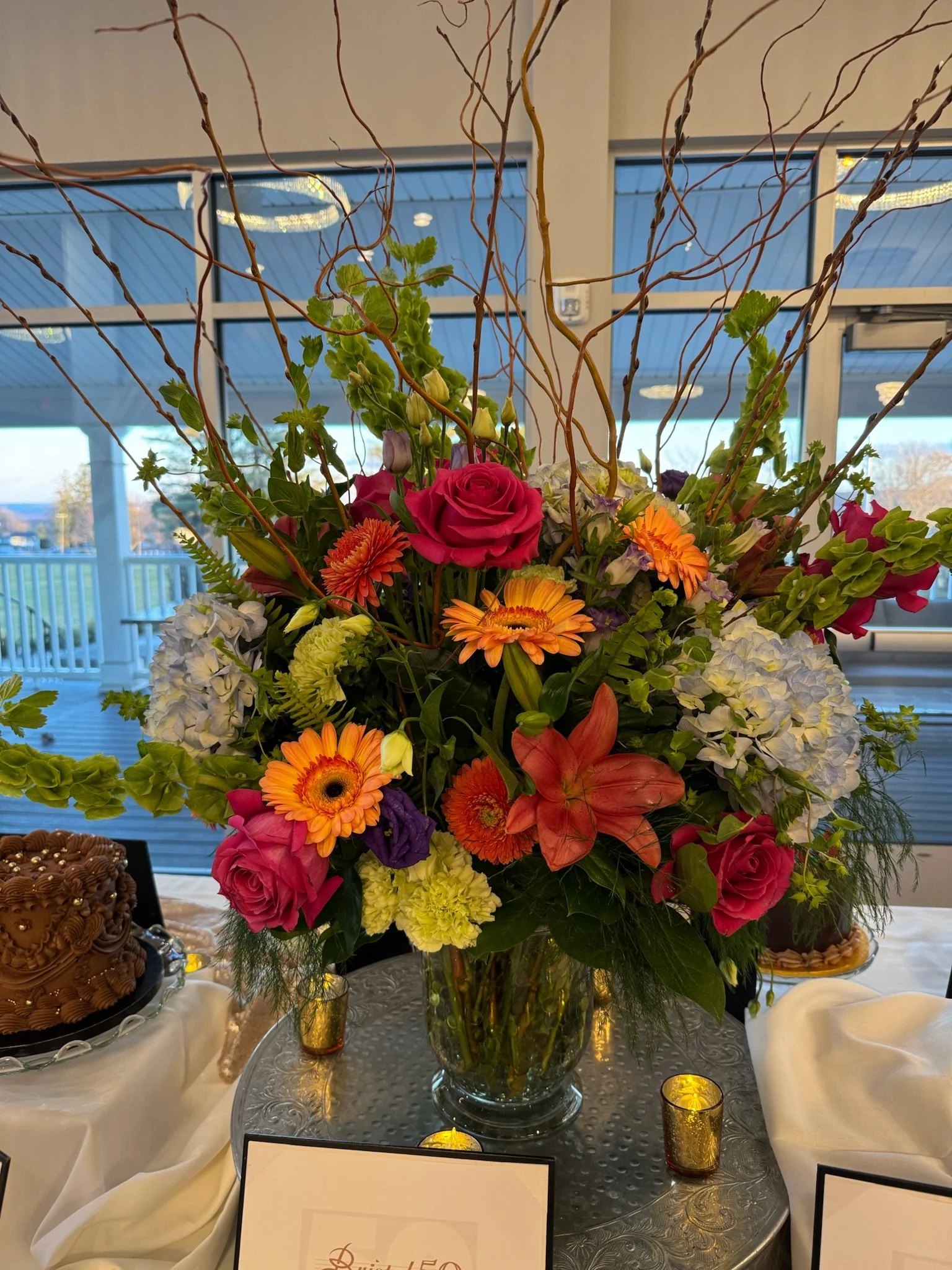 A large floral arrangement in a clear glass vase featuring pink roses, orange gerbera daisies, purple lisianthus, white hydrangeas, and green foliage, with twisting branches extending upward, displayed on a round table with a silver patterned surface