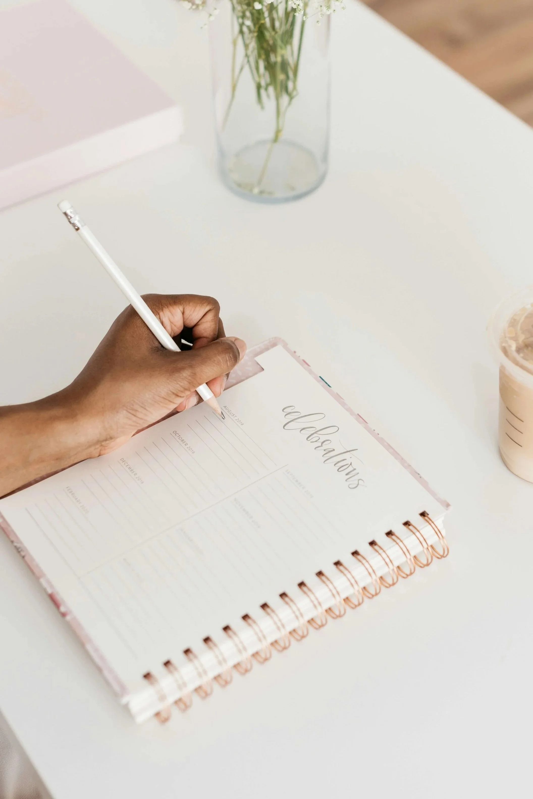 A person writing in a pink planner on a white desk, with a clear vase of flowers and a cup of iced coffee nearby.