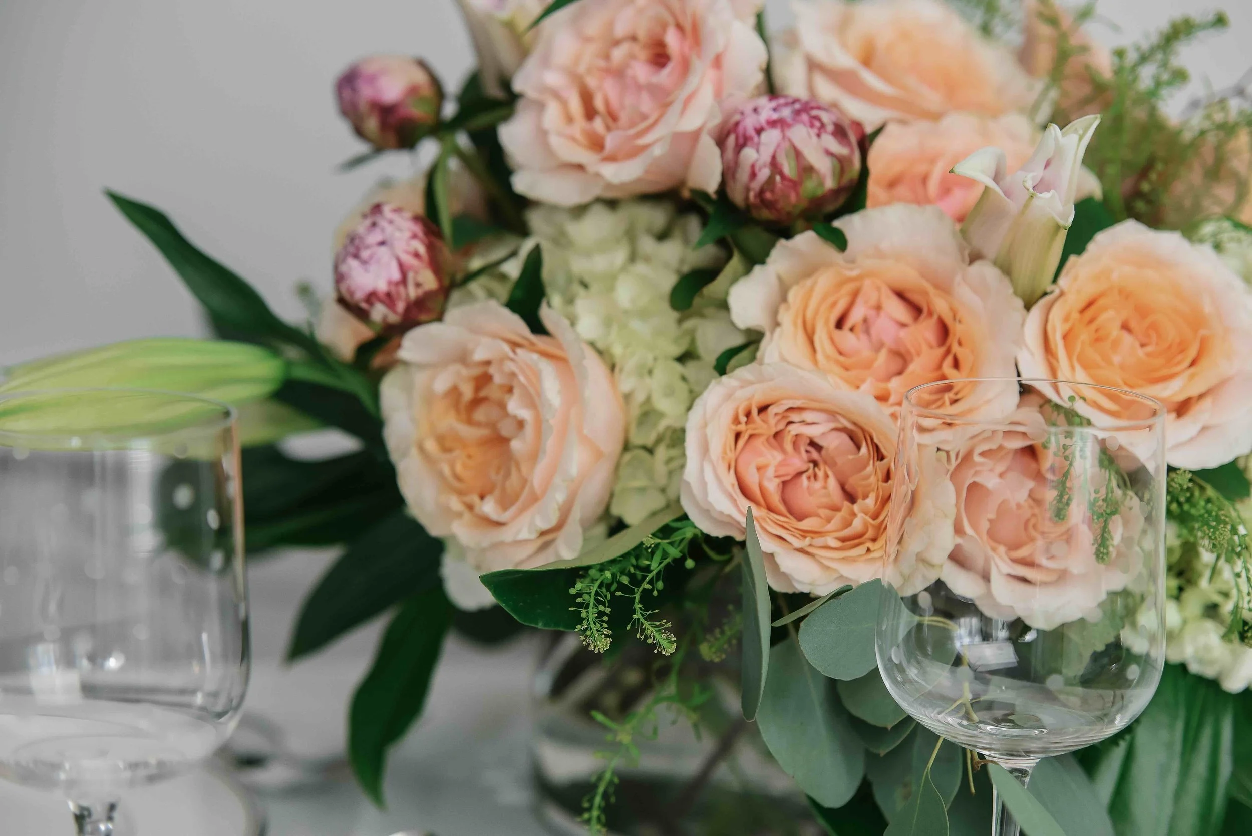 A bouquet of peach and pink roses, white flowers, and green foliage in a vase, with empty glassware in the foreground.