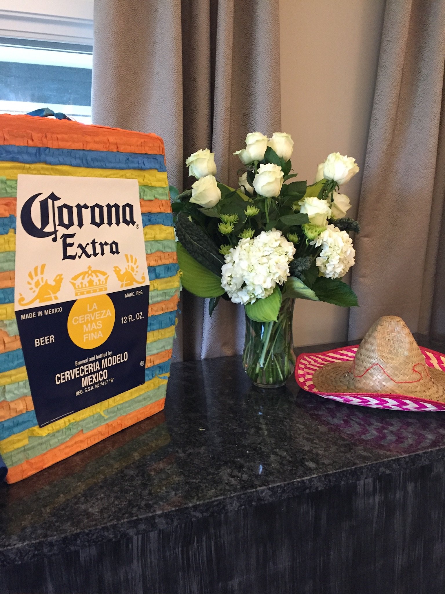 A colorful stack of multicolored paper napkins next to a small bouquet of white roses and green hydrangeas in a glass vase, on a dark granite countertop, with a straw sombrero on a pink and white woven mat nearby, and beige curtains in the background