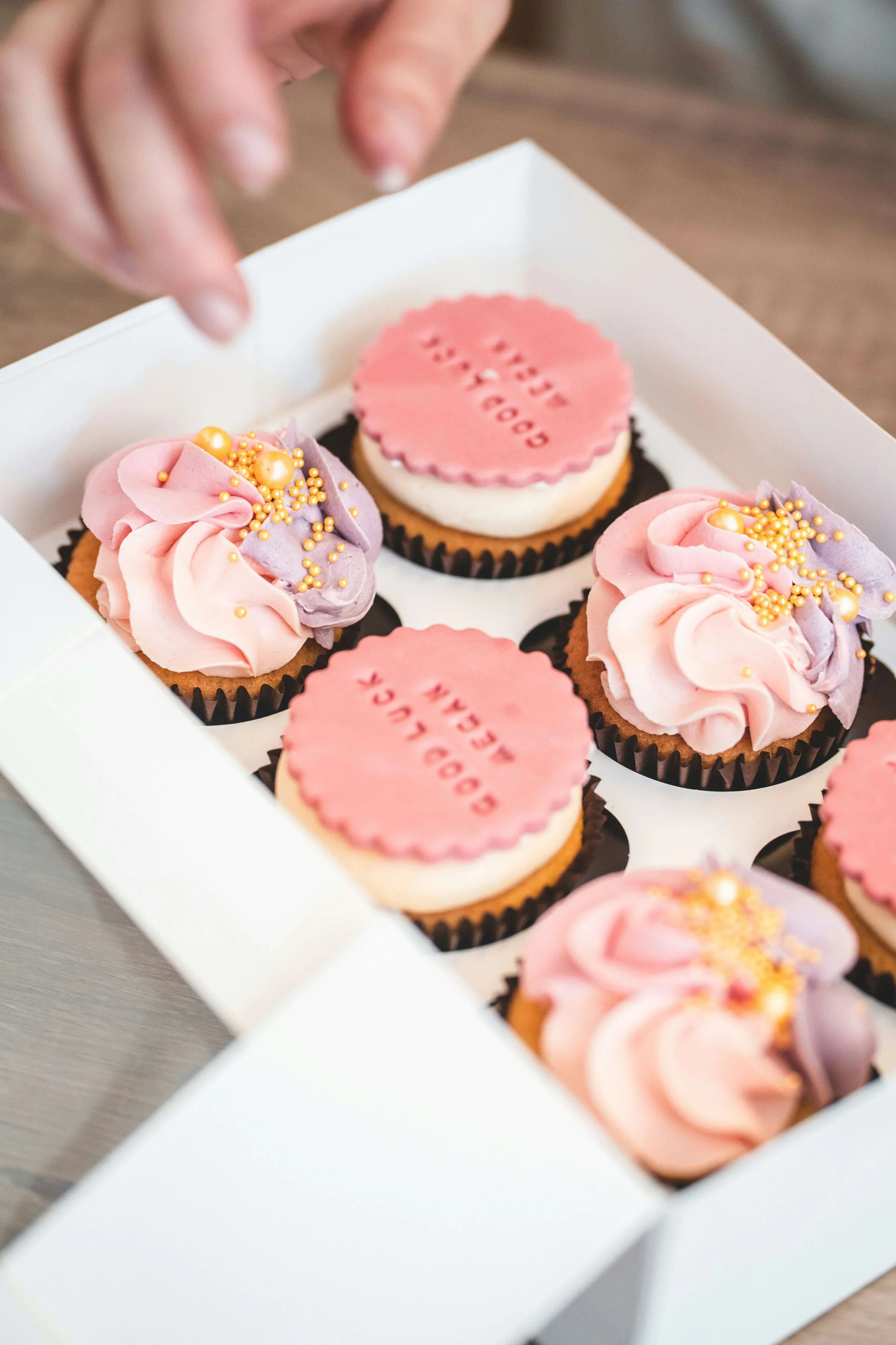 A box of assorted cupcakes with pink and purple frosting, decorated with gold and pearl sprinkles, and some featuring pink circular toppers with red text.