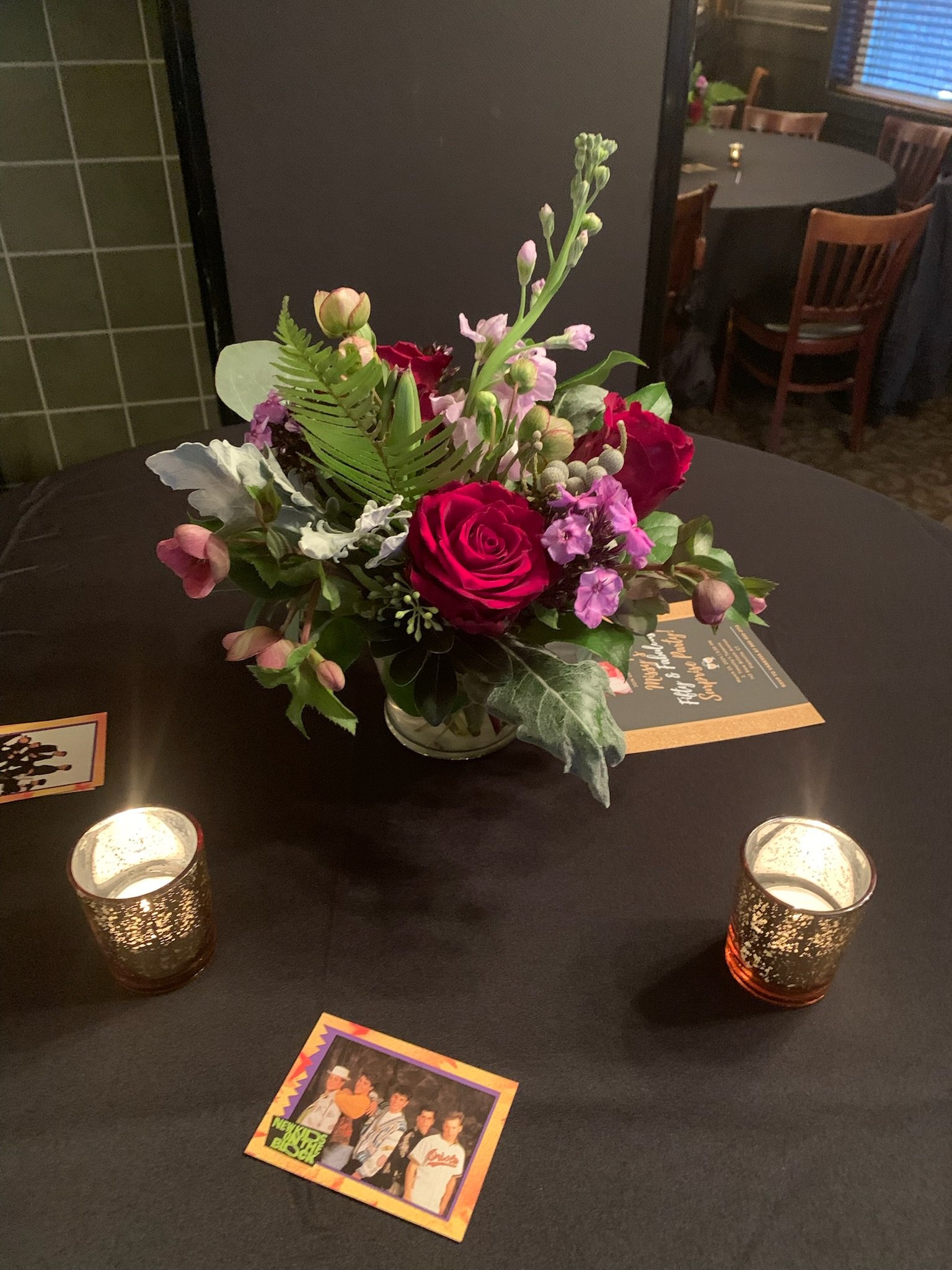 A table centerpiece with a bouquet of red, pink, and purple flowers and green foliage on a black table. Two lit candles in decorative holders are on the table, and a colorful magazine or flyer with a picture of a boy band and bright graphics is in th