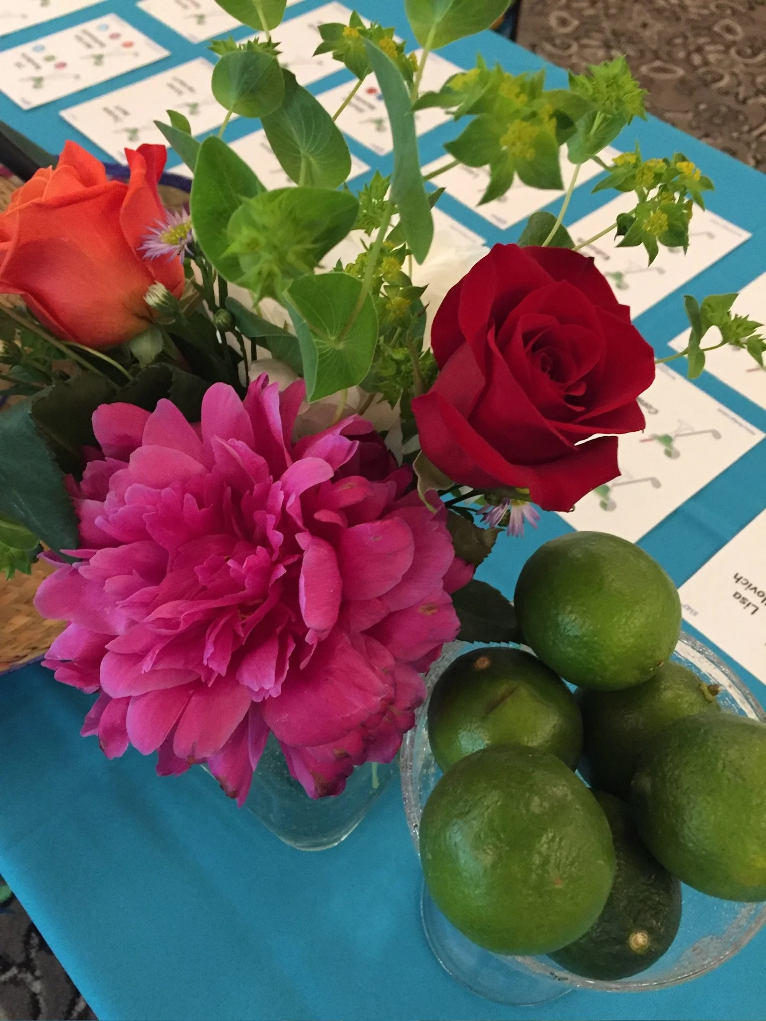 A glass bowl containing green limes next to a bouquet of colorful flowers including pink dahlias, orange roses, and small purple and yellow blossoms, set on a blue table with informational papers in the background.