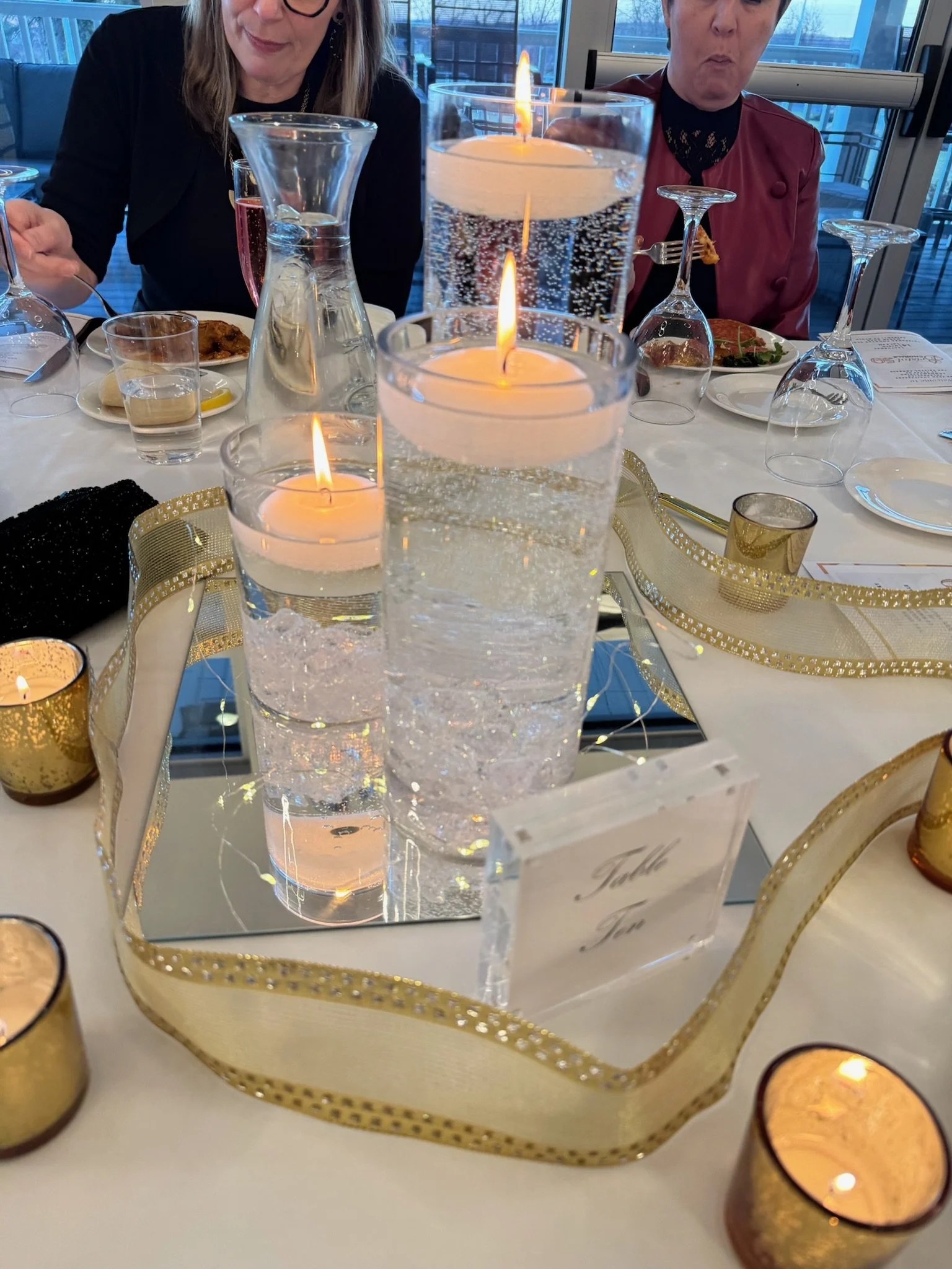 People sitting at a table with lit candles in glass holders, wine glasses, and plates of food, celebrating a special occasion indoors during the day.