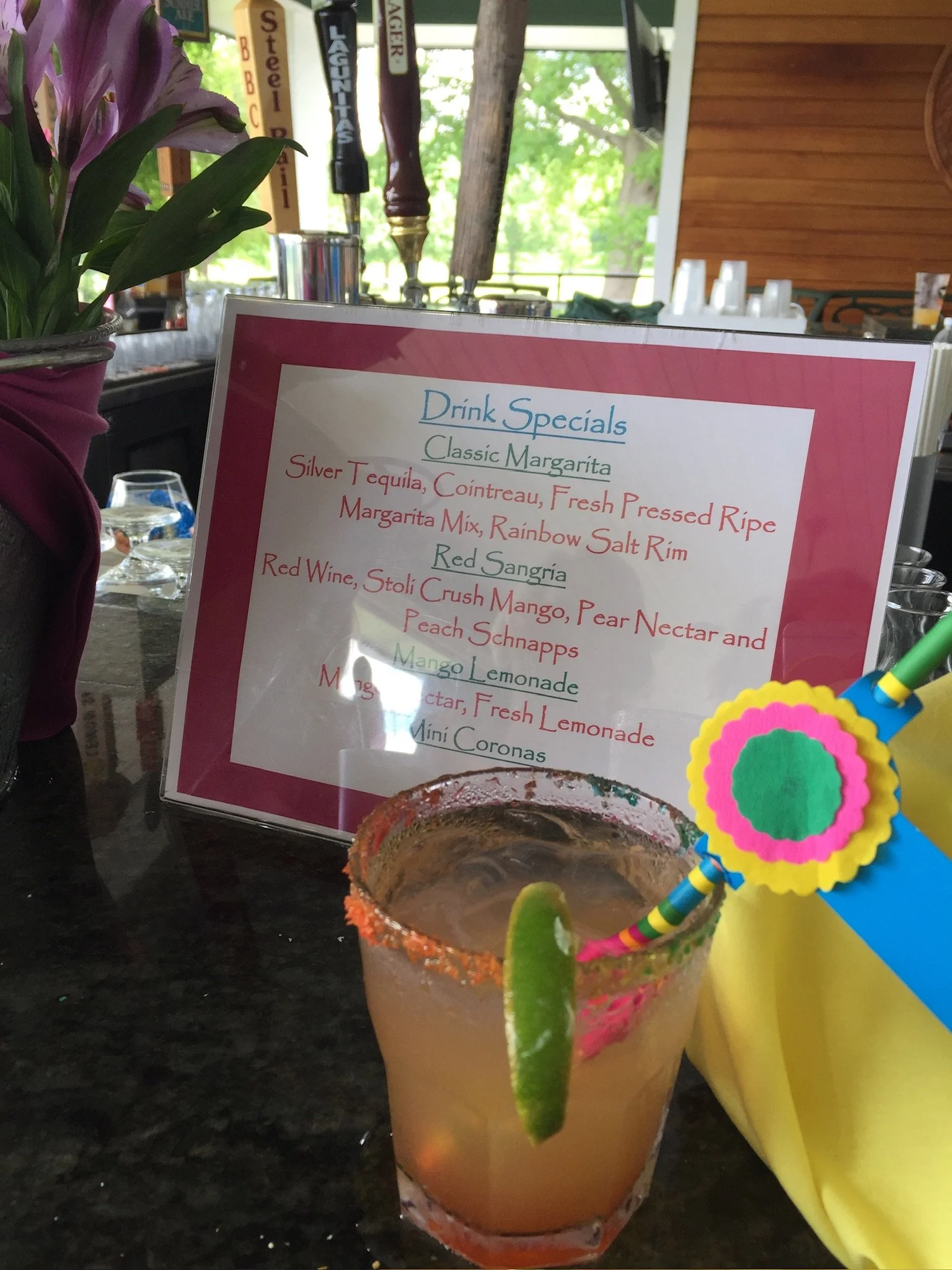A colorful Mexican cocktail drink garnished with a lime wedge and a slice of cucumber, inside a decorative glass, with a Fiesta-themed straw and paper decoration, on a dark table, with a menu and beer tap handles in the background.