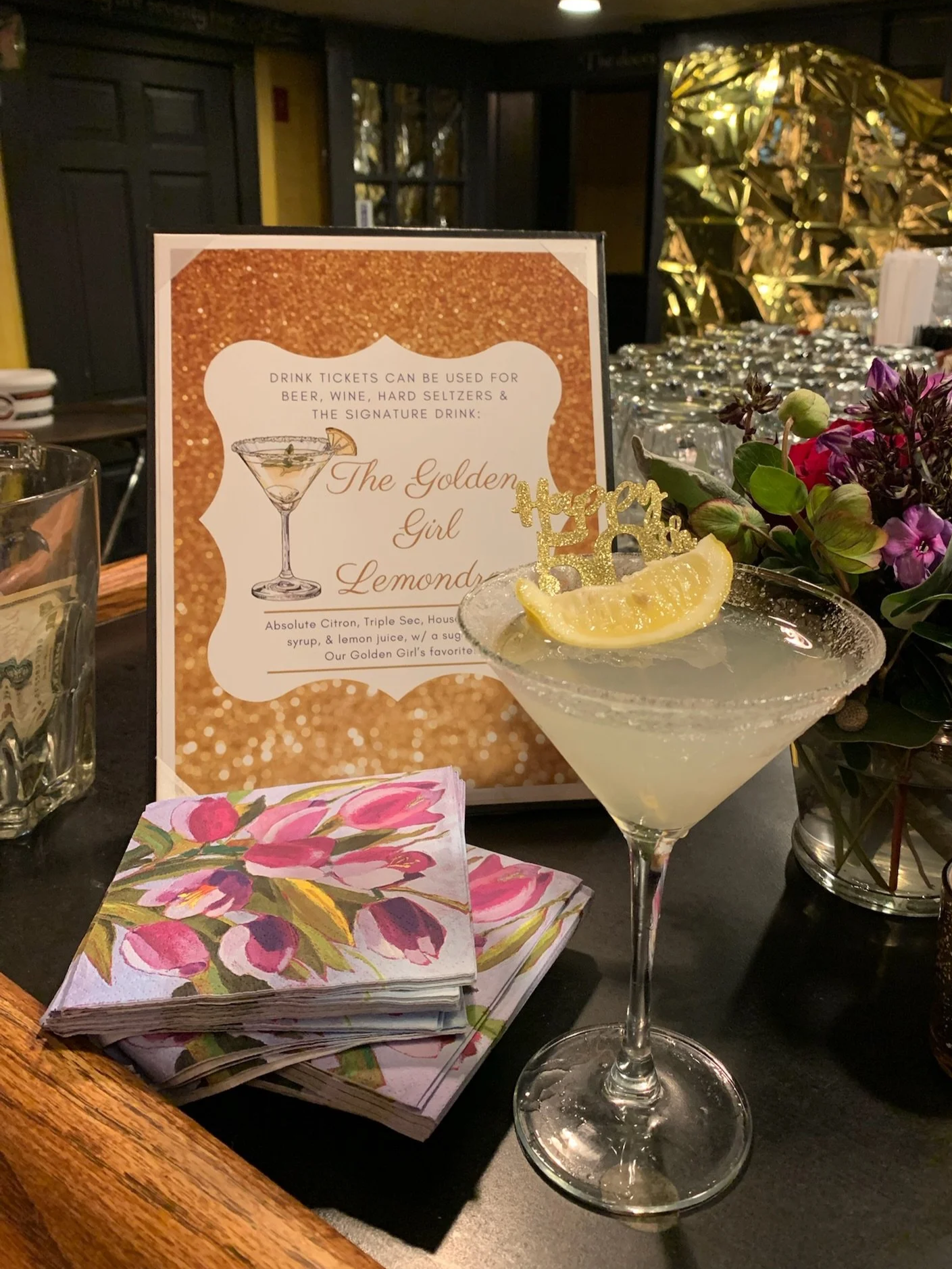 A bar or restaurant setting with a cocktail glass filled with a lemon and lime drink garnished with a slice of lemon and a decorative gold cake topper that says "Happy". There is a menu or sign in the background advertising a signature drink called "The Golden Girl" along with other drinks. On the table, there is a colorful floral napkin, a glass with some money inside, and a vase with purple and green flowers. The background is dimly lit with decorative gold foil or reflective material.