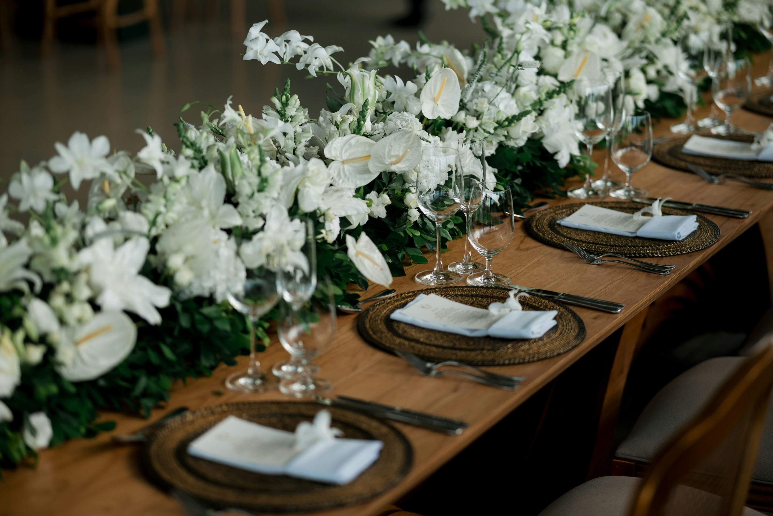 A long dining table decorated with a lush arrangement of white flowers and greenery, set with clear wine glasses, woven placemats, and neatly folded white napkins.