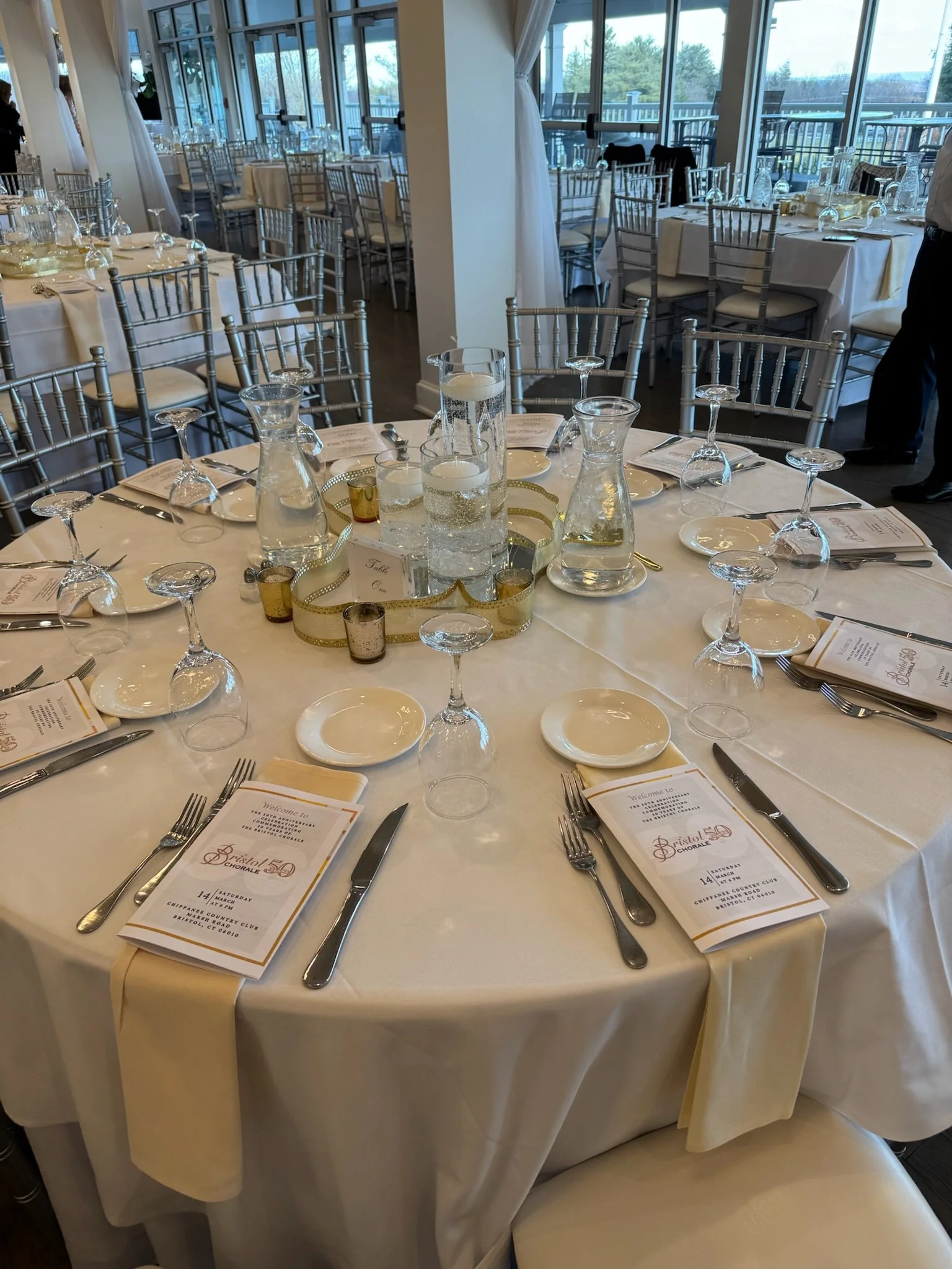 Round banquet table set with white tablecloth, place settings, glassware, menus, and decorative centerpiece in an event hall with large windows and empty chairs.