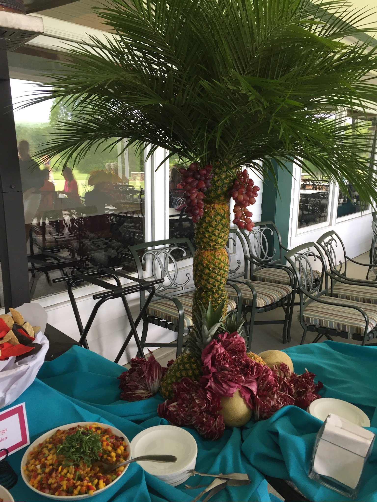 Decorative display of fresh pineapple with red grapes hanging in the middle, surrounded by flowers on a table with a blue cloth, with bowls of colorful salad and chairs in the background.
