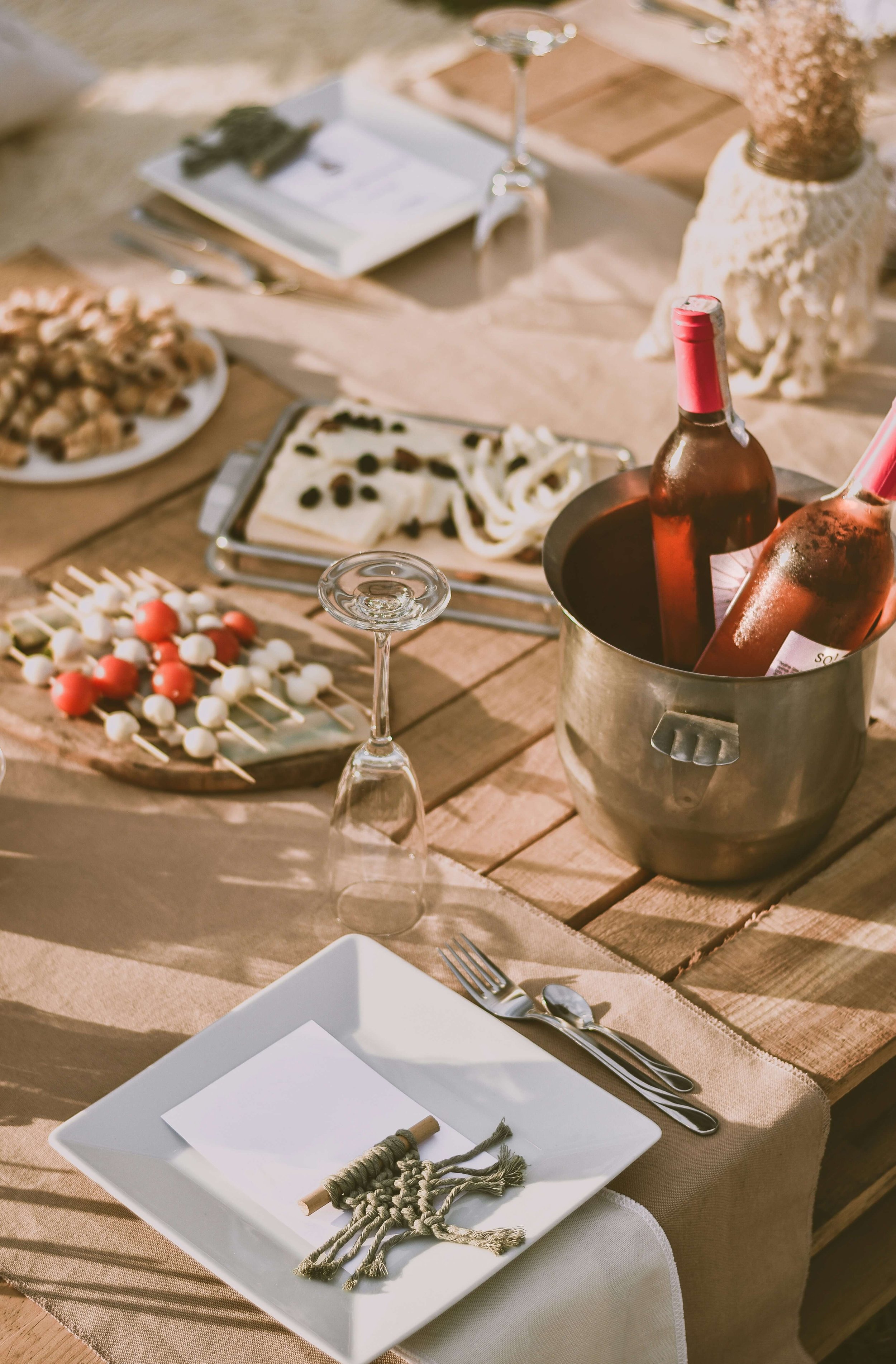 A table set for a meal with a white square plate, a fork, and a spoon, with a beige table runner. The table has various snacks including cheese, olives, cherry tomatoes, and nuts, along with a bottle of rosé wine in an ice bucket, an empty wine glass, a menu, and some decorative items in the background.