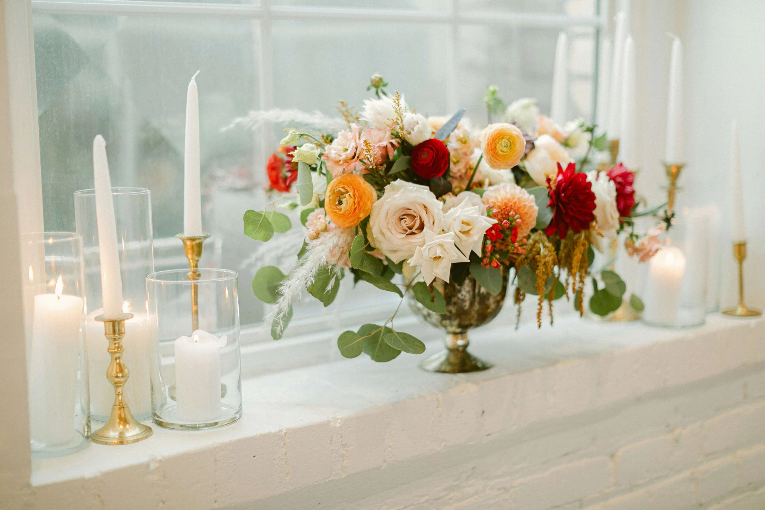 A floral arrangement with pink, white, red, and orange flowers and green leaves in a decorative vase on a window sill, accompanied by white candles and glass candle holders.