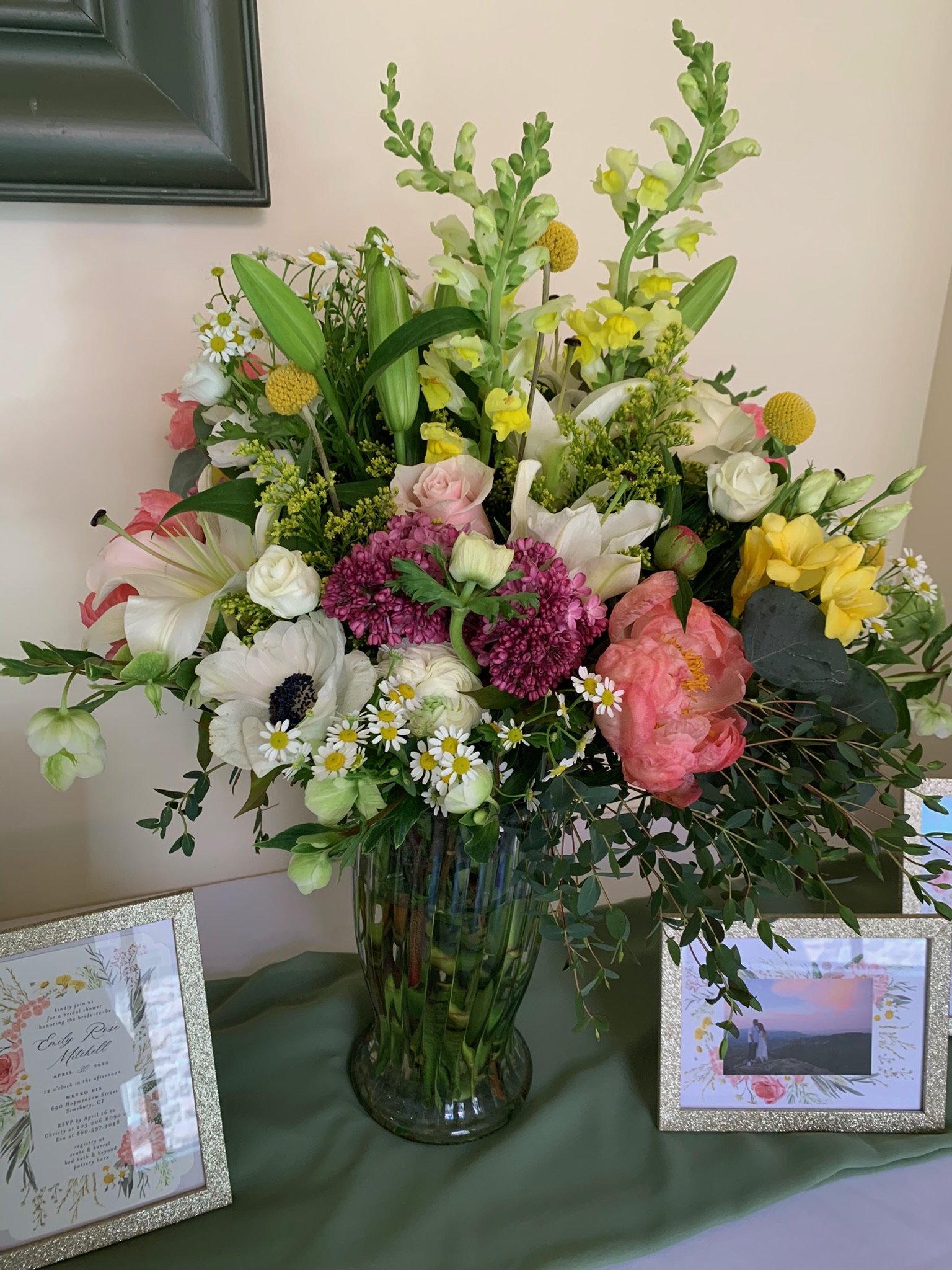 A large glass vase filled with a colorful bouquet of various flowers and greenery displayed on a table, standing next to framed pictures and an invitation or program sheet.