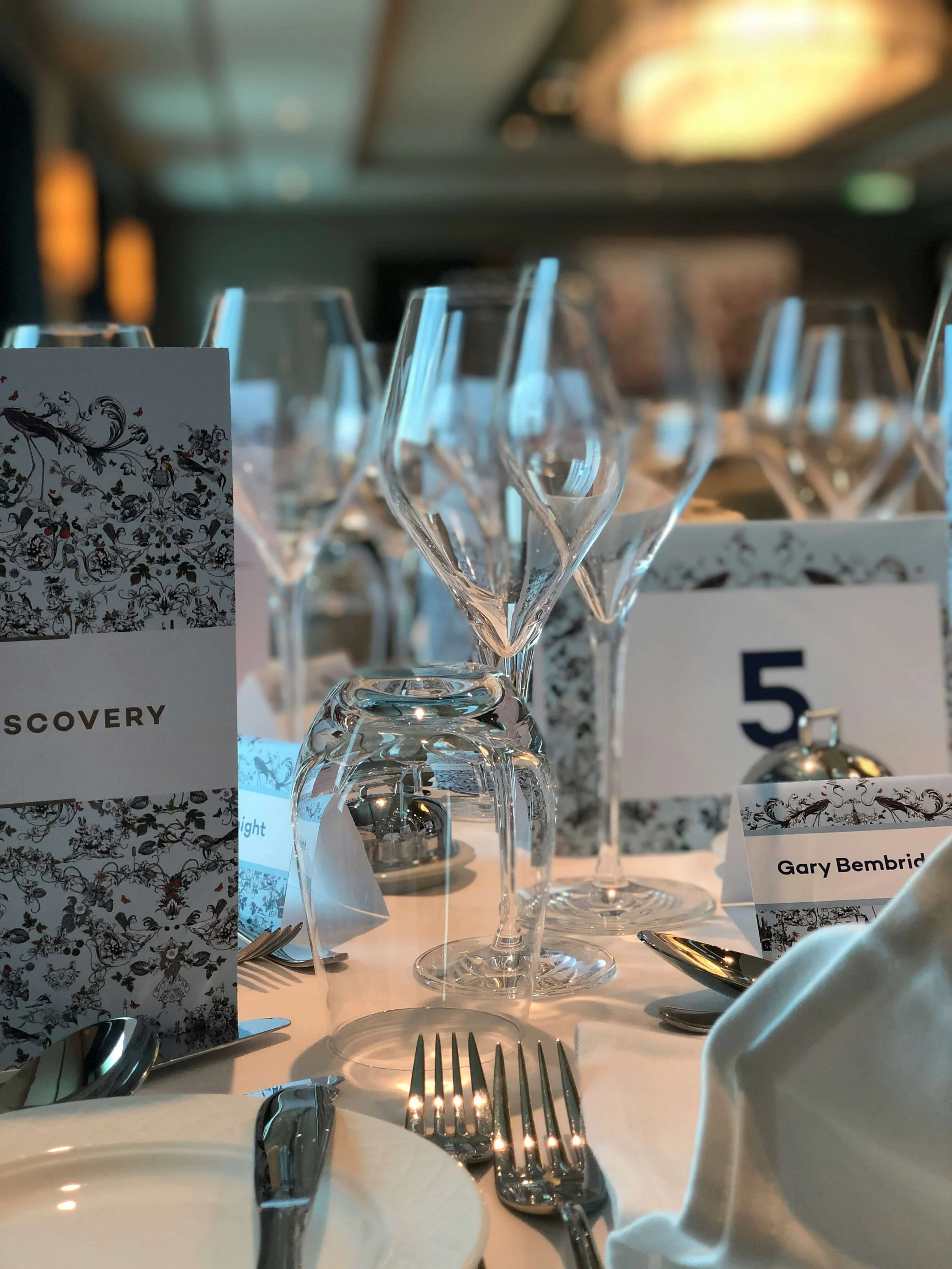 A formal dining table set with multiple upside-down wine glasses, silverware including forks and spoons, and place cards with the names Gary Bembridge and Discovery. The table is decorated with a white tablecloth, and there is a decorative card with a floral pattern in the background. The setting appears to be in a banquet or conference room with blurred background lights.