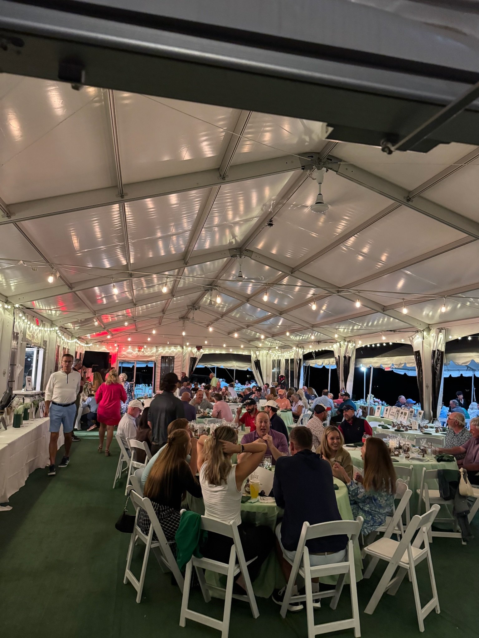 People seated at long tables inside a large decorated tent, attending a social event or celebration, with some standing and chatting, illuminated by strings of hanging lights.