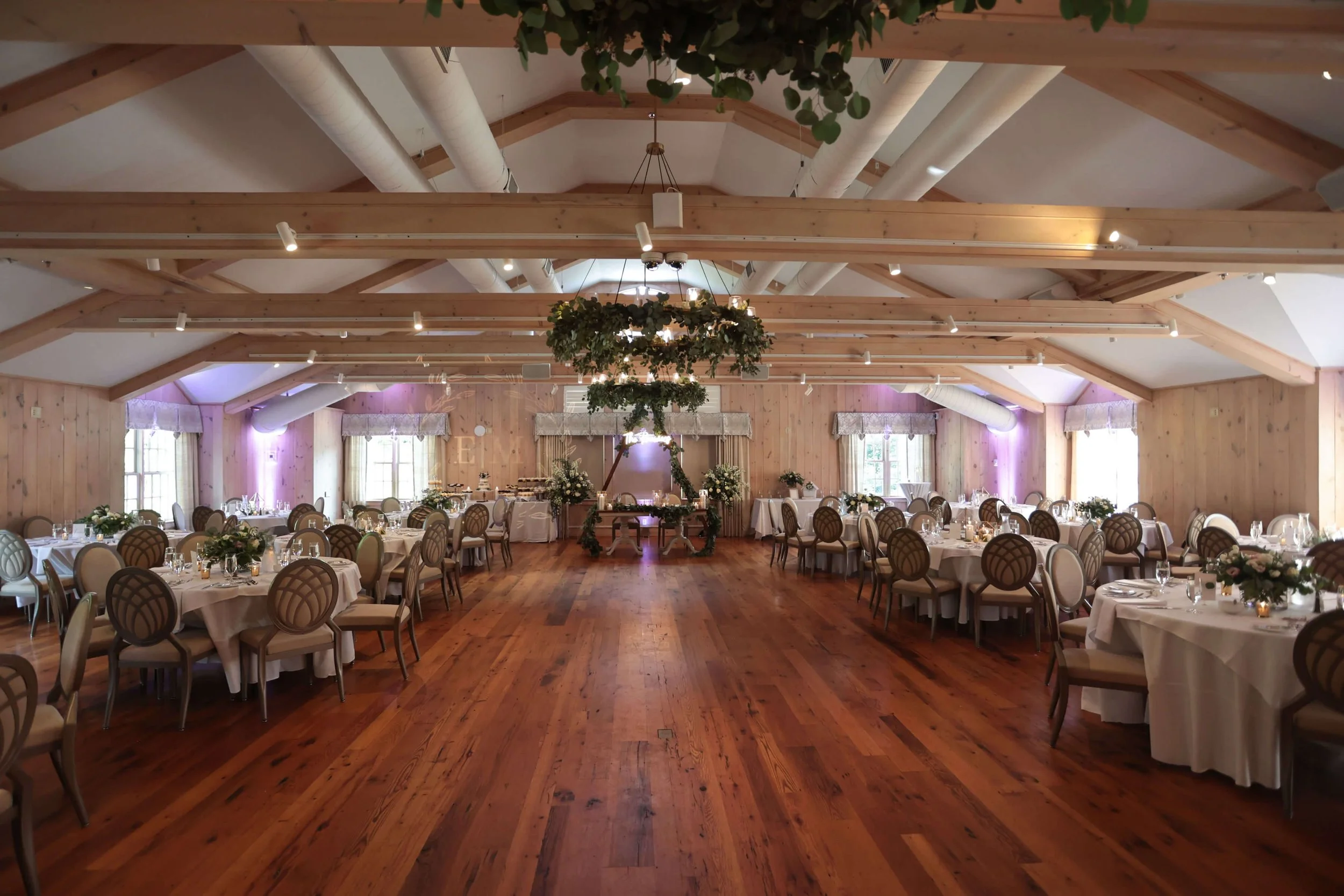 A spacious banquet hall decorated for an event with round tables covered in white tablecloths, surrounded by elegant chairs. The hall has wooden floors, wood-paneled walls, and a high vaulted ceiling with exposed beams and large white air ducts. Overhead lighting and purple ambient lights illuminate the space. A floral arrangement and candles are in the center of the room, suggesting a wedding or celebration.