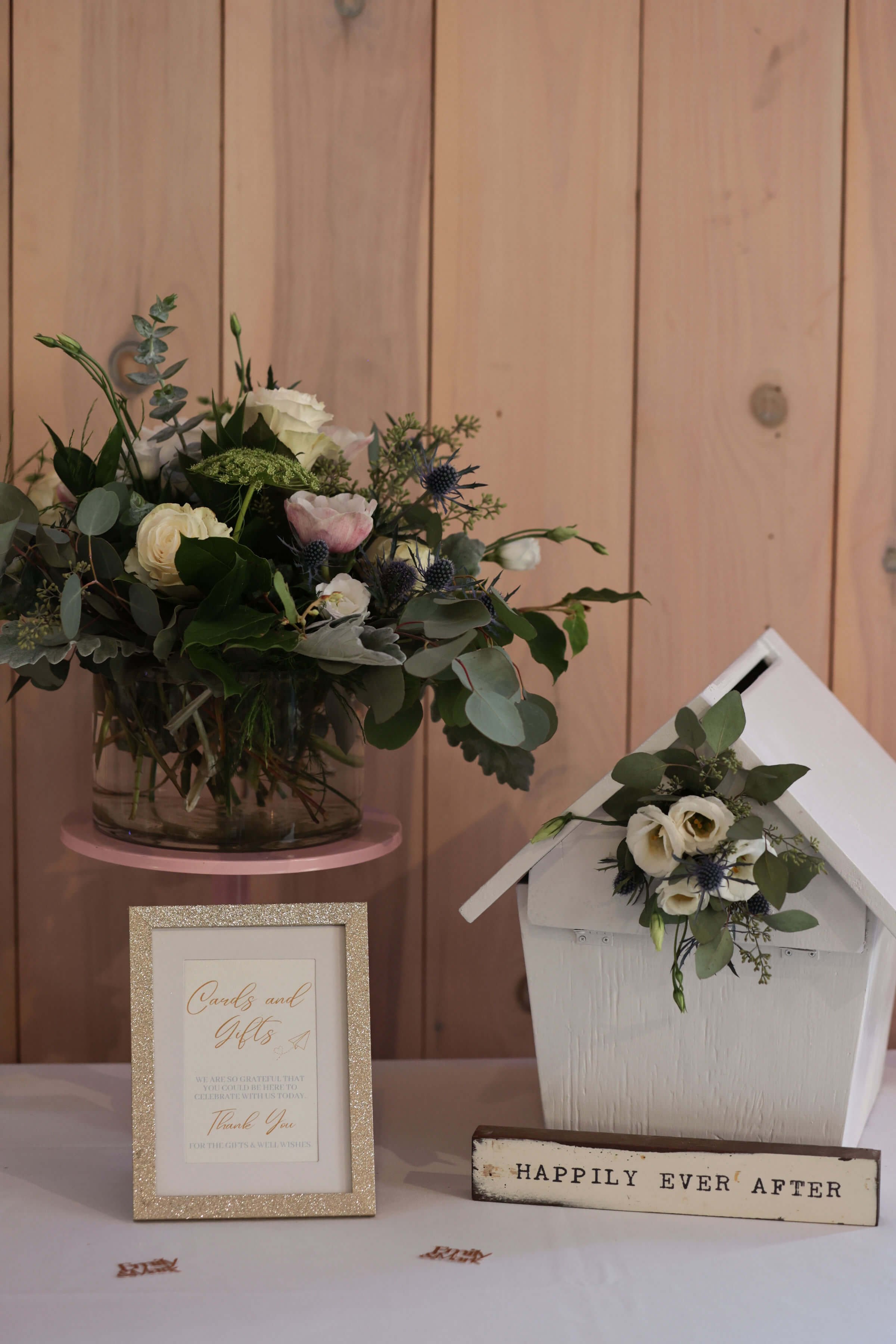 A floral arrangement with white, pink, and purple flowers and green leaves in a glass vase, set on a pink round stand. Next to it is a white container with similar flowers, and there are signs reading 'Cards & Gifts' and 'Happily Ever After' on the table, with a plain wooden wall background.