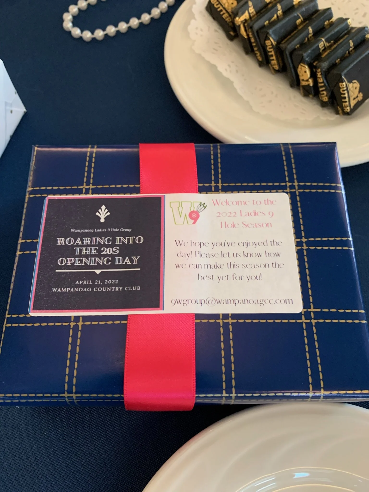 A blue gift box with a red ribbon, a white doily, a string of pearls, and a white dish with wrapped chocolates on it, all placed on a dark tablecloth.