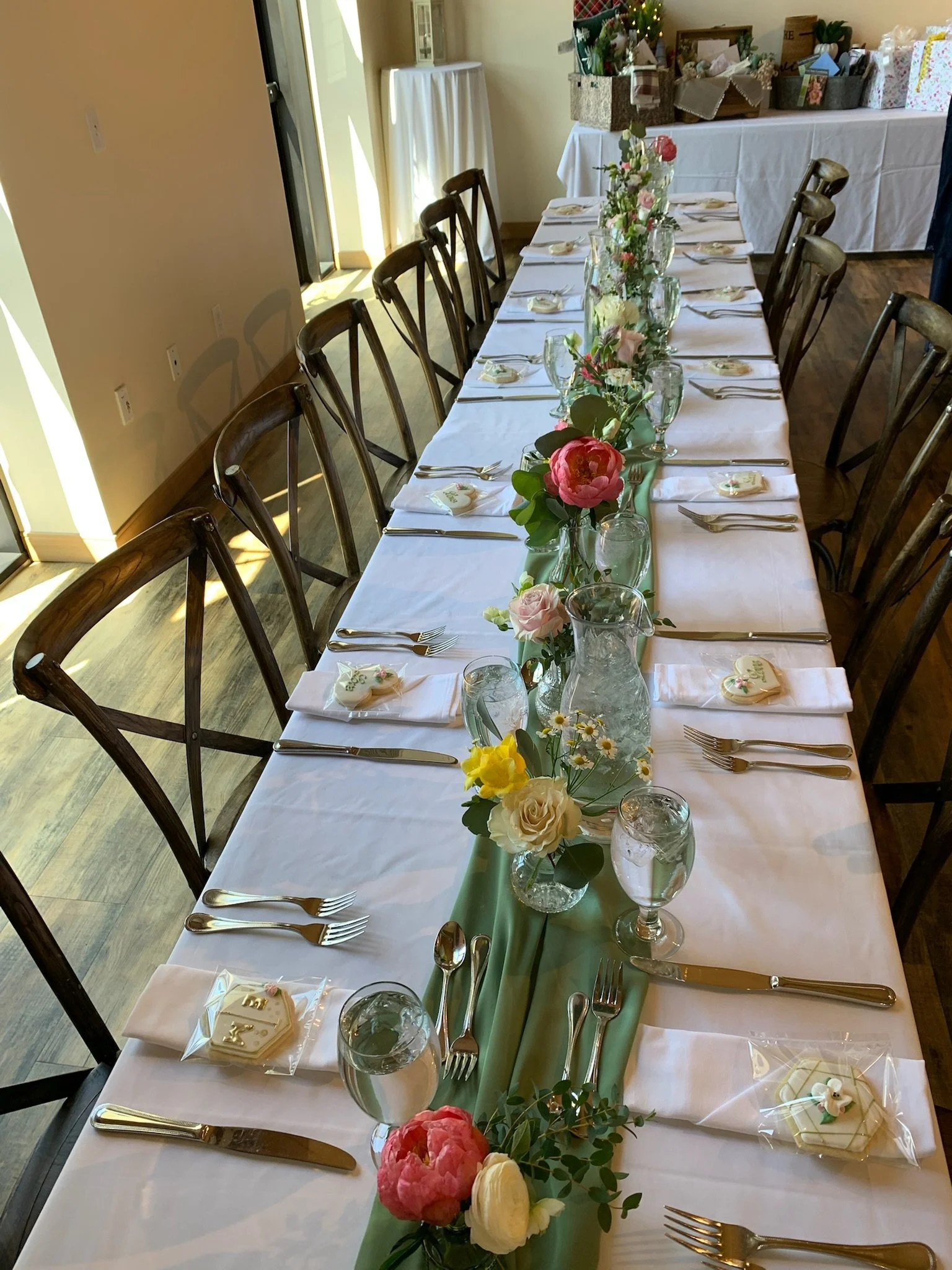 A long rectangular dining table set for a formal event with white tablecloths, gold utensils, glassware, and small decorated cookies at each place setting. A green runner with floral arrangements in vases running down the center of the table. In the background, there are gift boxes and wrapped presents.