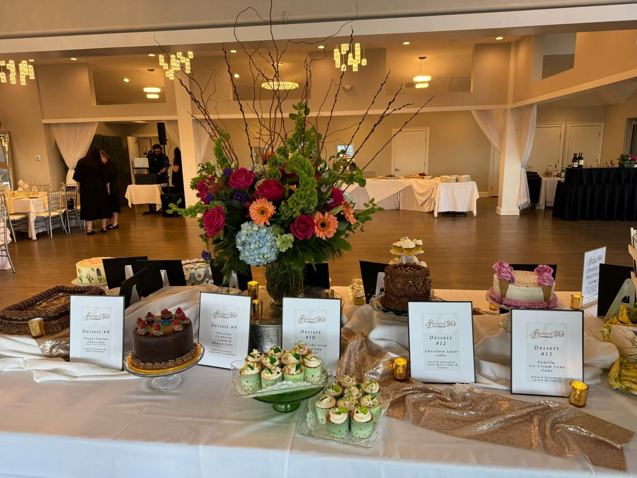 A large floral arrangement with pink, purple, orange, and green flowers on a table, with cakes and dessert labels in front. The background shows a banquet hall with tables, chairs, and people setting up.