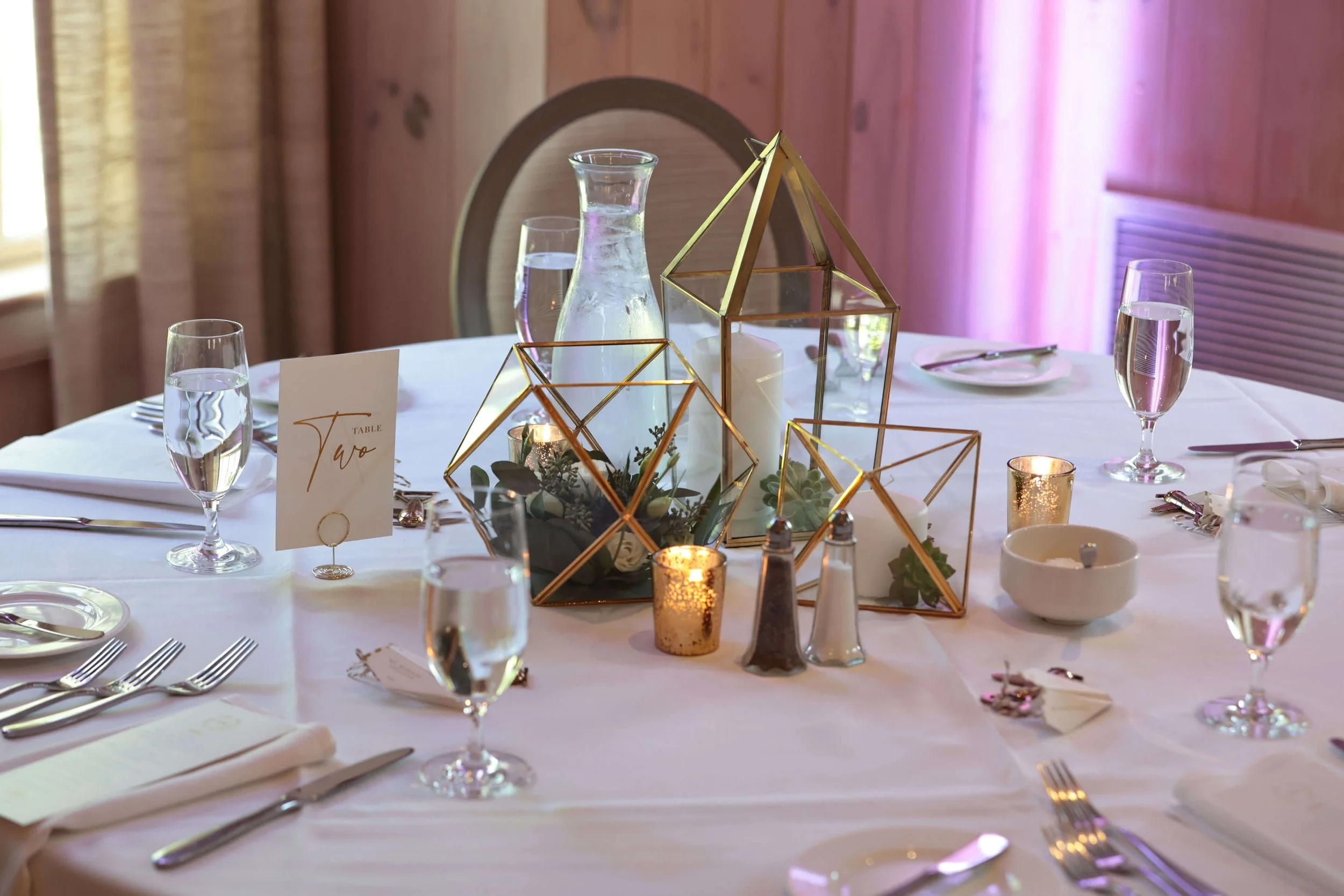 A round banquet table decorated with glass candle holders, gold geometric glass terrariums, and glass vases with water, set for a formal event, with a table number card labeled "Two".