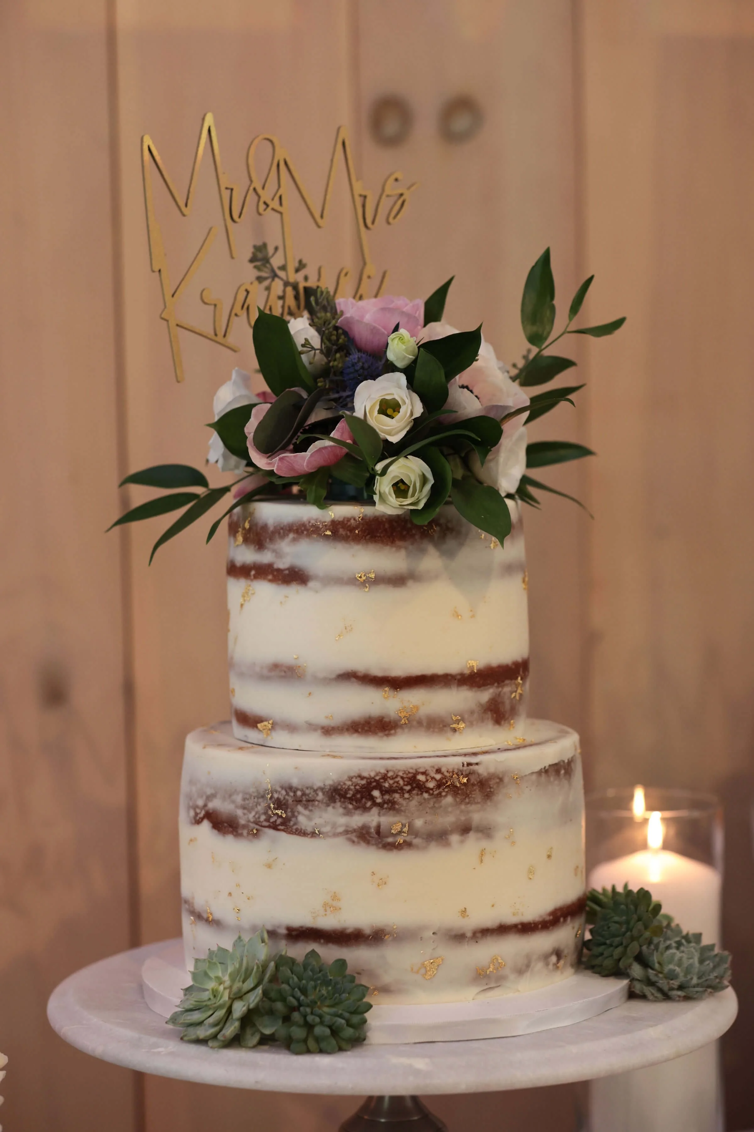 A two-tiered semi-naked wedding cake decorated with white, pink, and purple flowers, green foliage, and gold leaf accents, topped with a gold cake topper that reads 'Mr & Mrs'. The cake is placed on a white marble cake stand, and there are greenery decorations at the base.