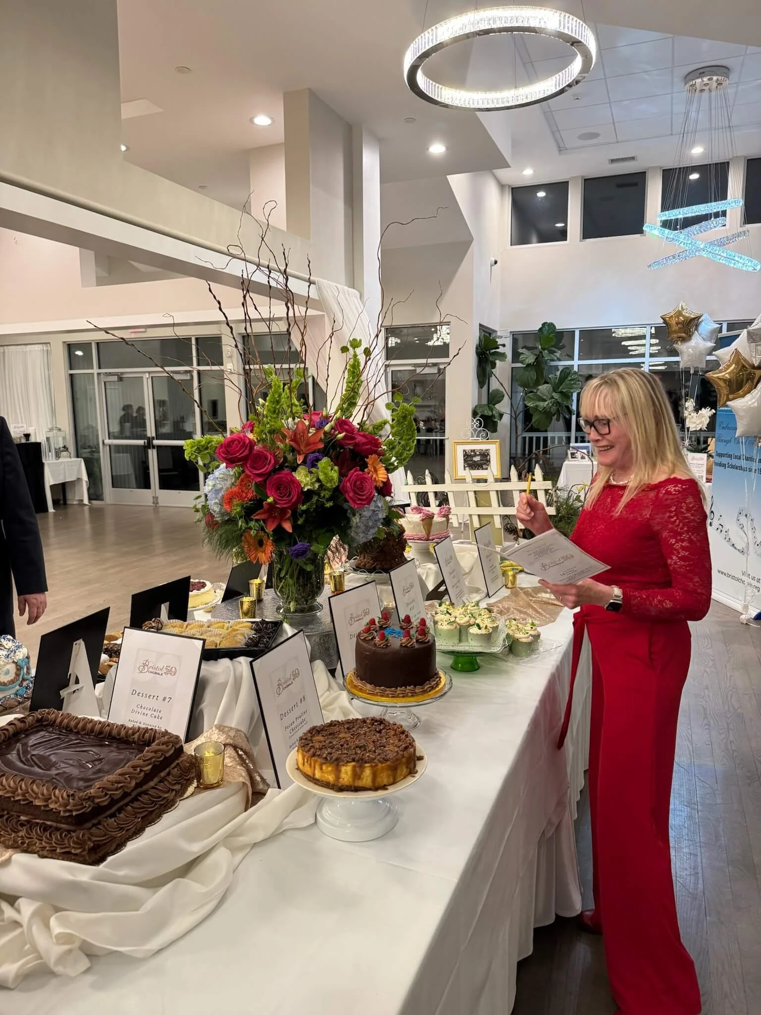 A woman in a red dress and glasses standing at a dessert table with an assortment of cakes and treats, in a modern, well-lit event space decorated with balloons and floral arrangements.