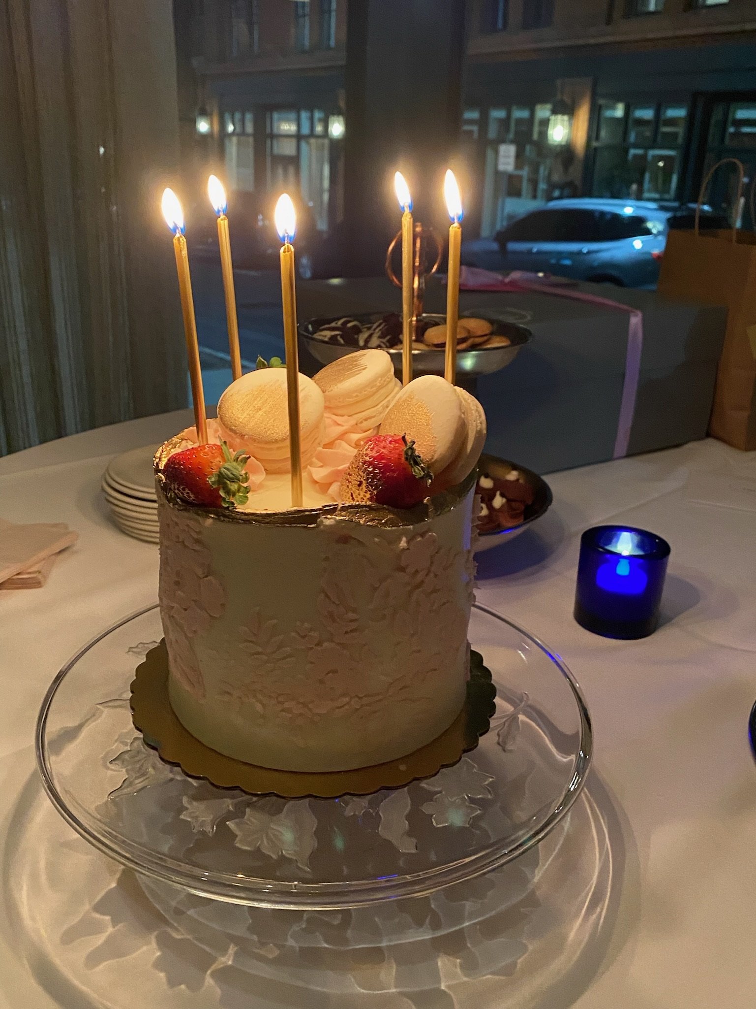 A birthday cake with five lit candles, decorated with strawberries, macarons, and cream, placed on a glass cake stand. In the background, there is a bowl of cookies and a  stack of plates on a table, with a window showing a parked car outside.