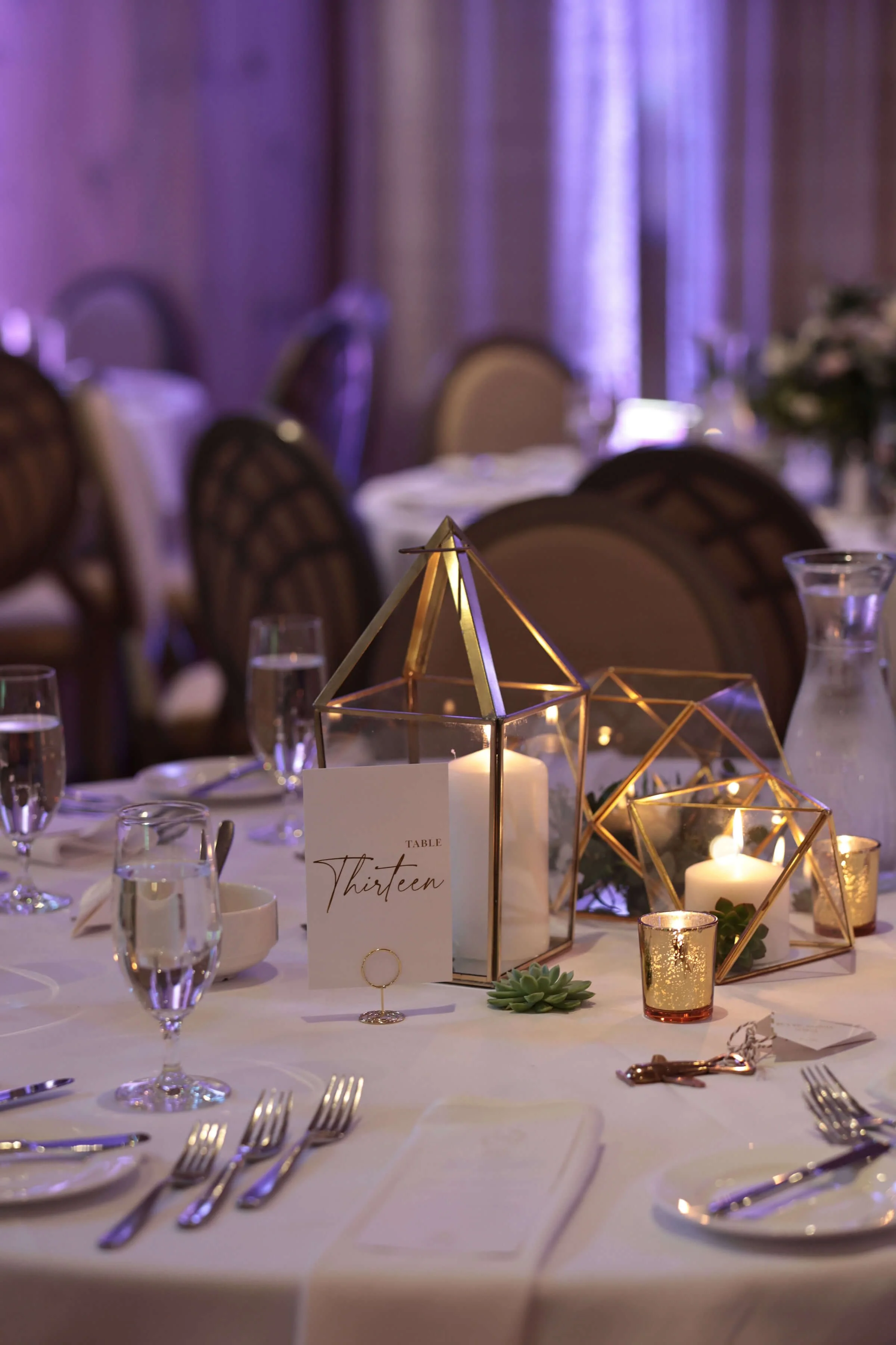 Close-up of a decorated banquet table with gold geometric candle holders, candles, small succulents, glassware, and a table number sign reading 'Thirteen' in elegant script, set for an event with purple and blue lighting.