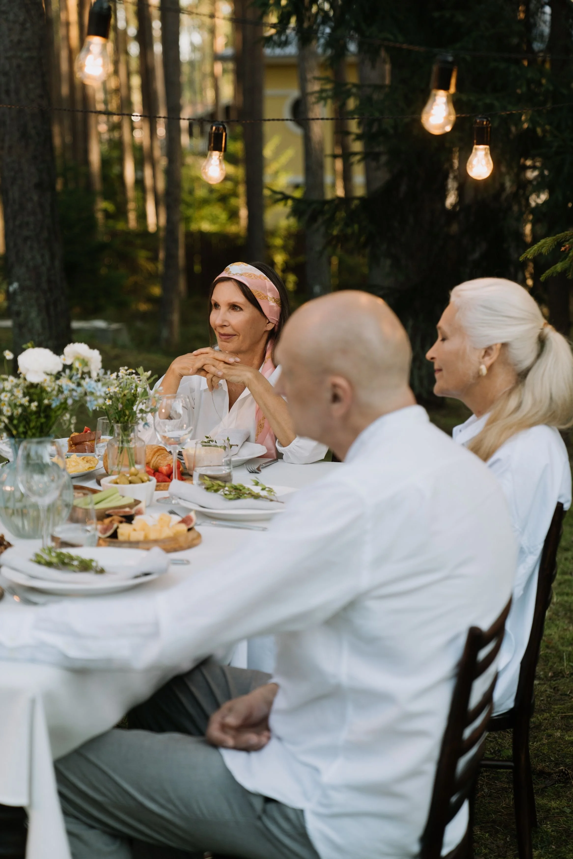 People sitting at a long outdoor dining table in a wooded area, decorated with string lights and flowers, enjoying a meal.