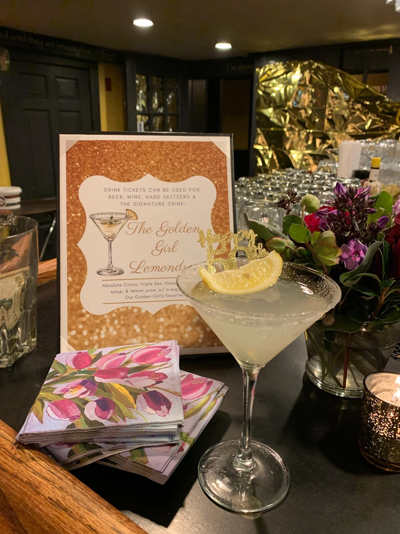 A cocktail glass with a lemon wedge garnishing the rim and a lemon slice at the base of the glass, placed on a black surface with a floral napkin, a printed menu, a bouquet of purple flowers, and various bar items in the background.