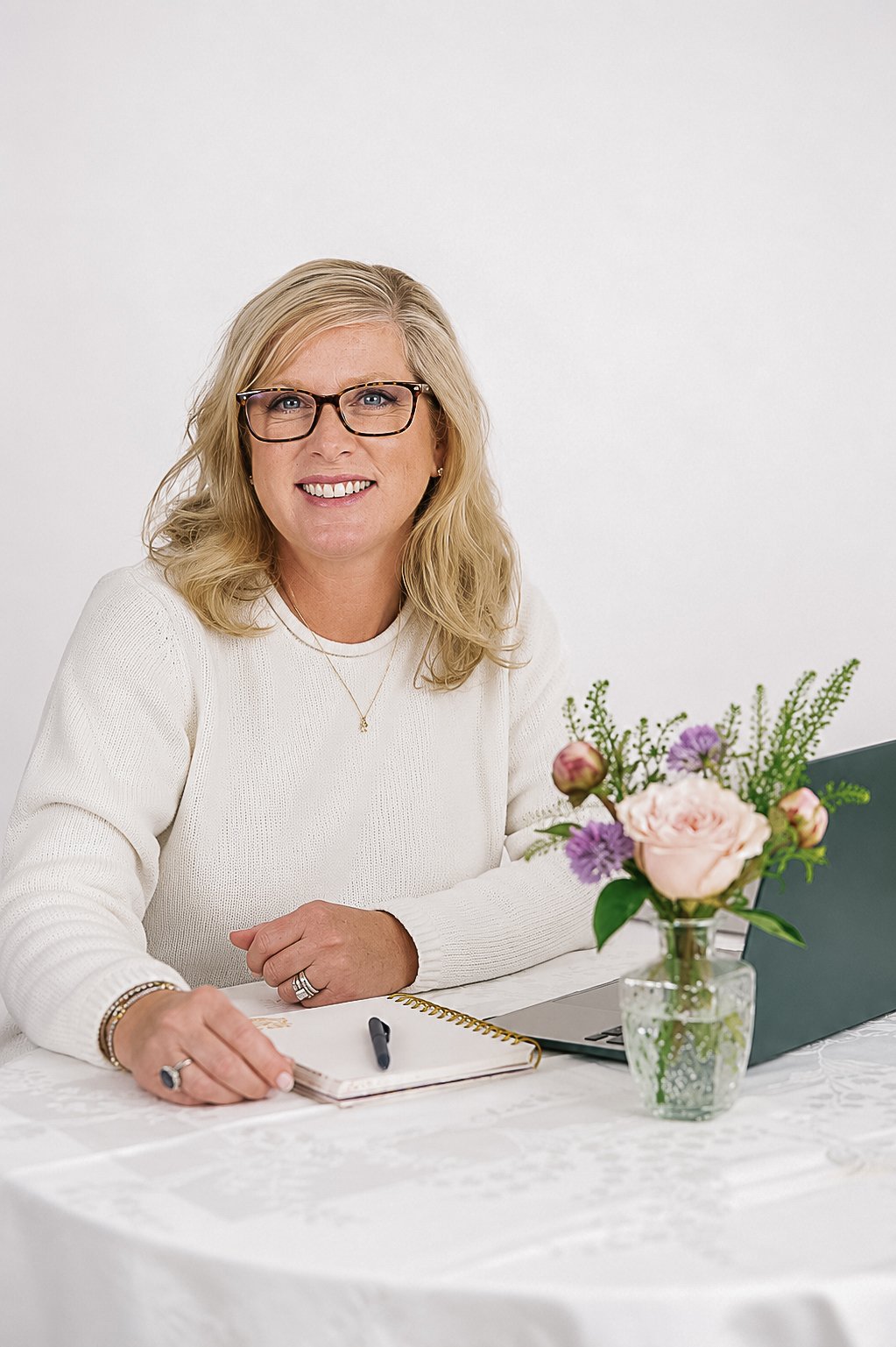 A smiling woman with blonde hair, glasses, and a white sweater sitting at a table with a notebook, pen, laptop, and a vase of flowers, in front of a white background.