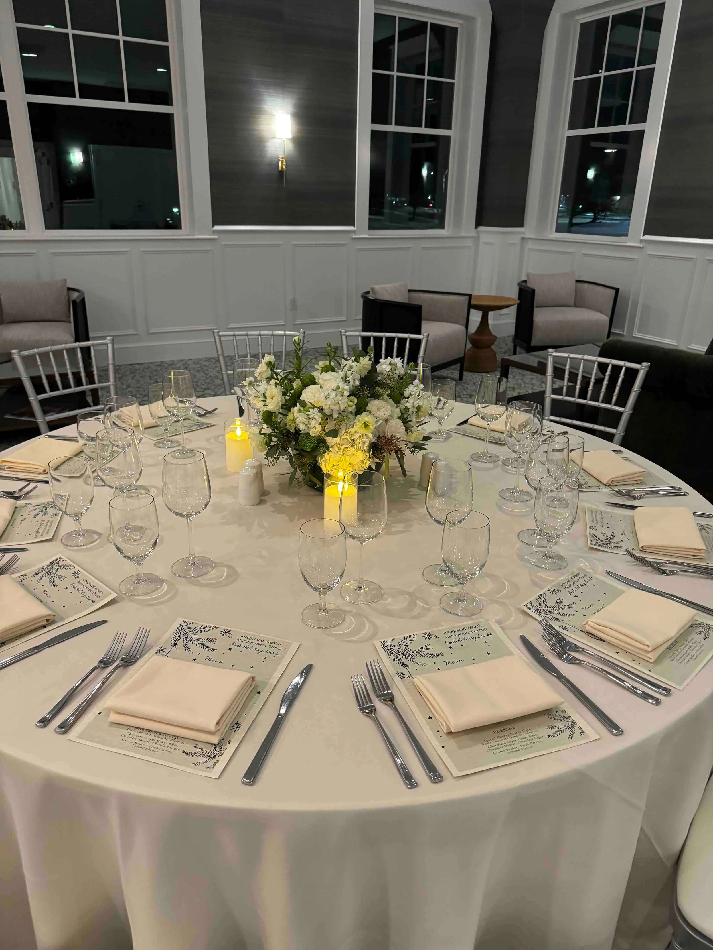 A round banquet table set for a formal event with a white tablecloth, multiple wine glasses, and beige napkins. The table features a floral centerpiece with white and green flowers and yellow candles. Menus or place cards are placed at each seat. The background shows a room with large windows, cushioned chairs, and a dark wall with a wall-mounted light.