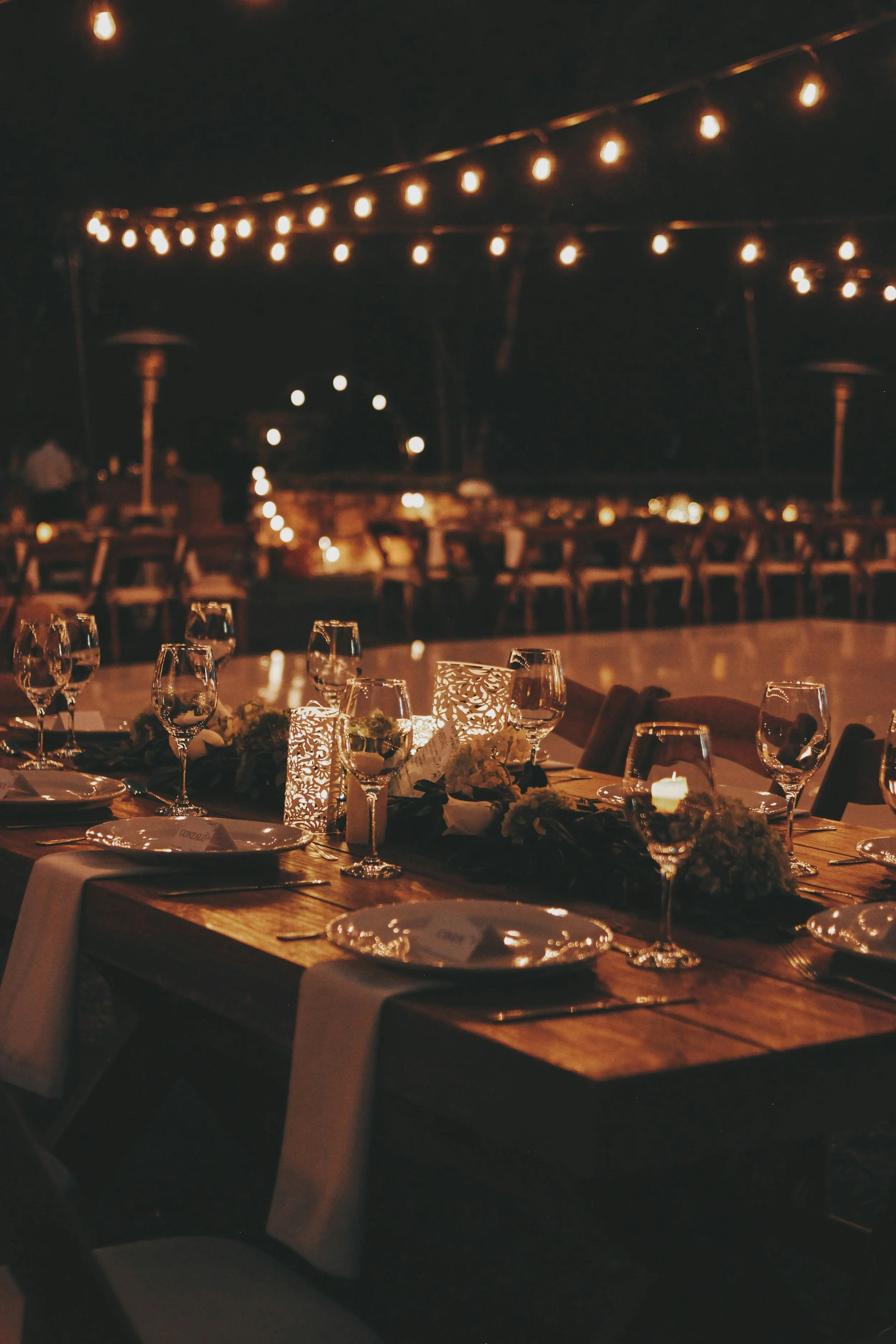 A nighttime scene of an outdoor dining area decorated with string lights overhead, with tables set with wine glasses, candles, and floral centerpieces.