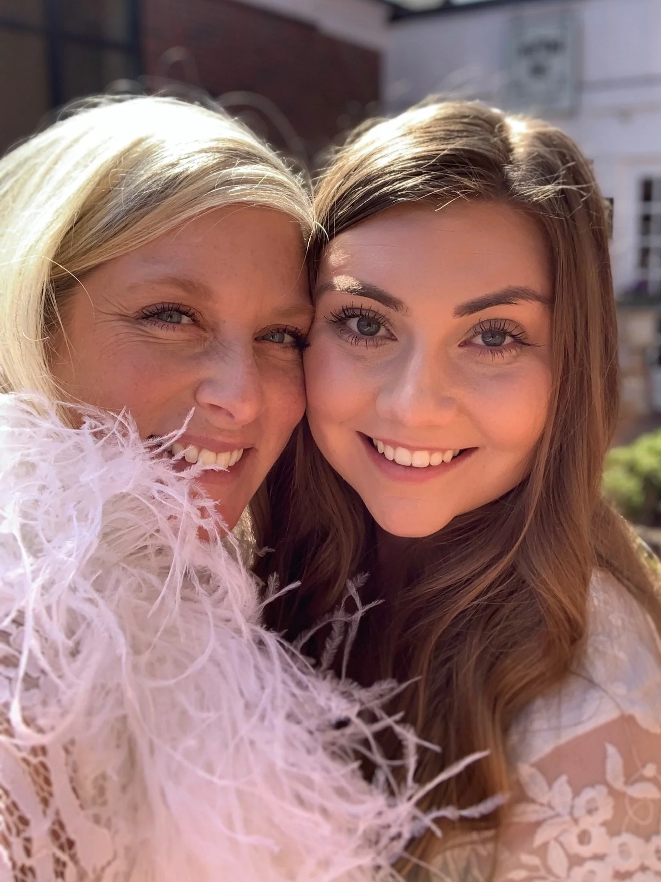 Two smiling women in close-up, one with blonde hair and the other with brown hair, posing outside in sunlight. The blonde woman is wearing a pink, fluffy feathered garment, and they are both smiling at the camera.