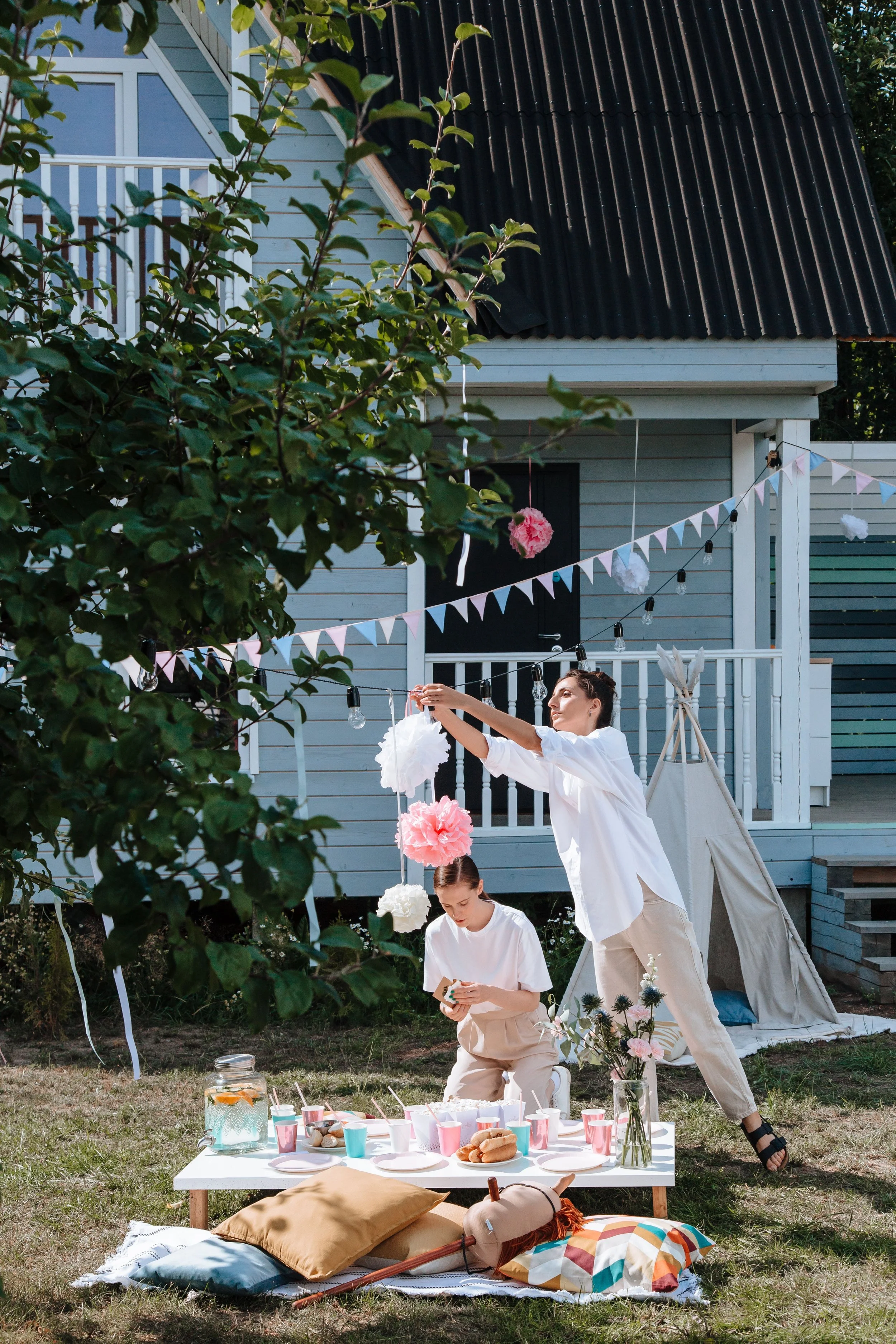 People having a backyard party with a table of food, drinks, and balloons, while a woman adjusts decorations on a string of hanging paper pom-poms and banners.