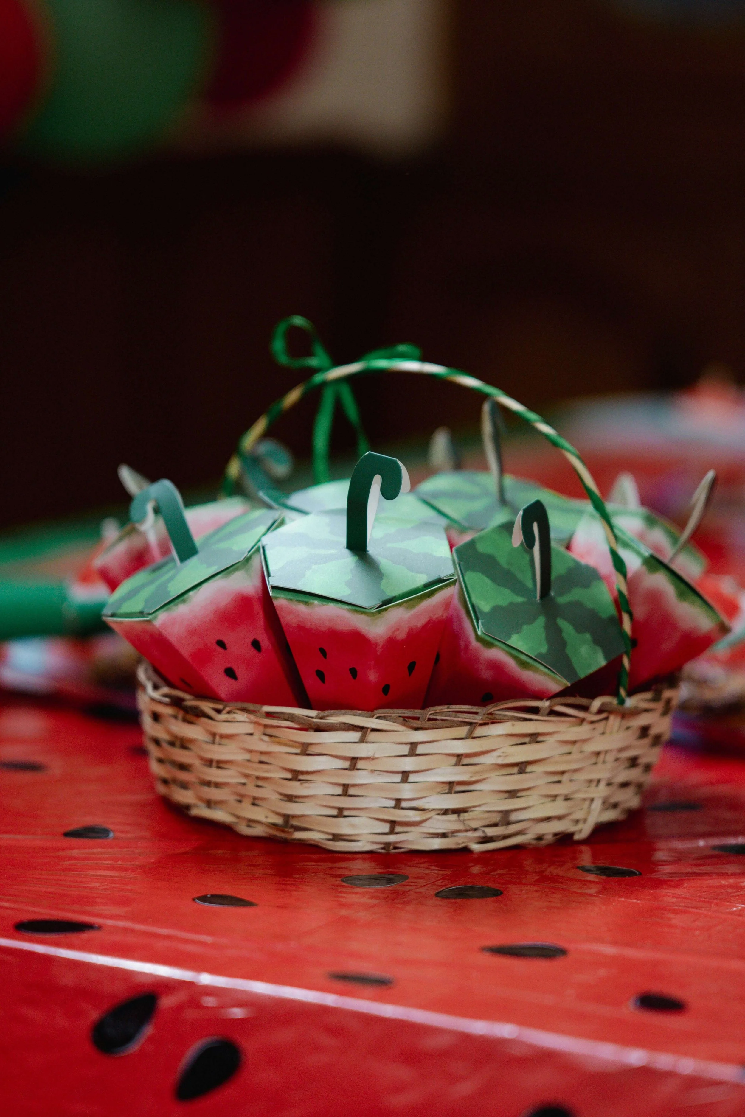 A basket of small, watermelon-shaped paper crafts with green, white, and pink colors, set on a glossy red surface with black ink splatters.