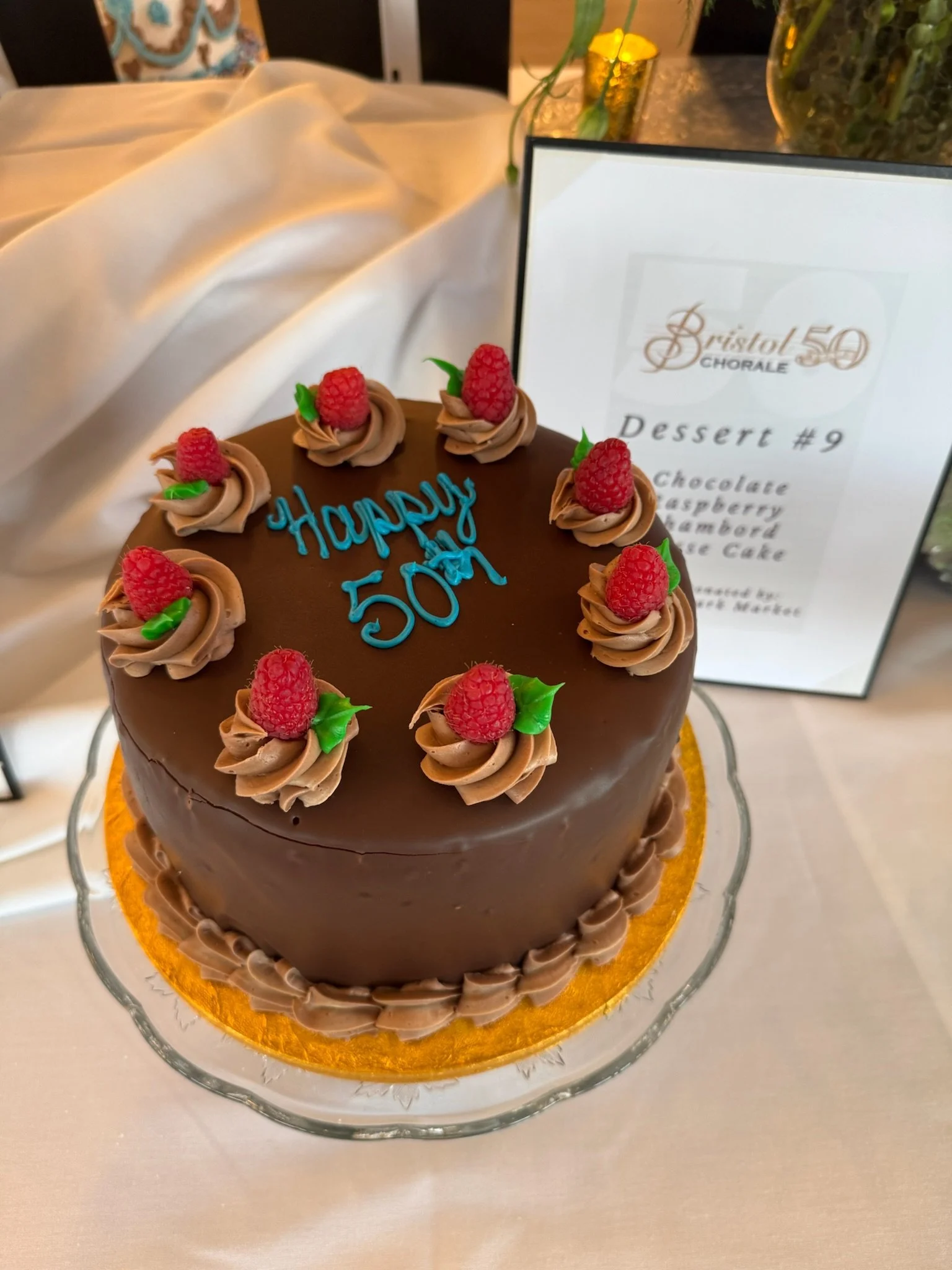A chocolate cake decorated with raspberry and green leaf toppings, with blue icing text reading 'Happy 50th' on top, next to a sign with a menu for a 'Bristol 50' choir event.