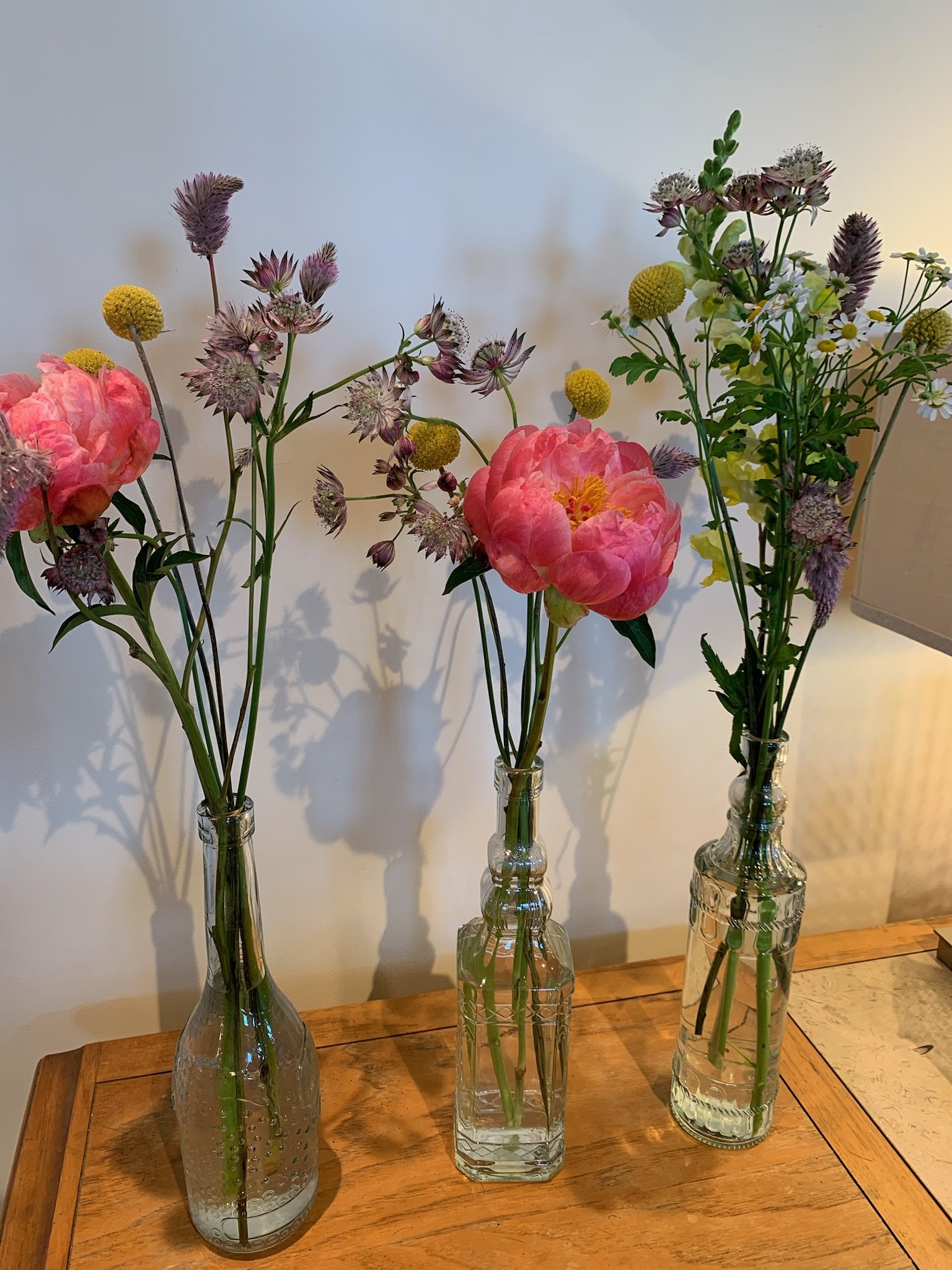 Three glass vases with different sets of flowers on a wooden table against a plain wall.