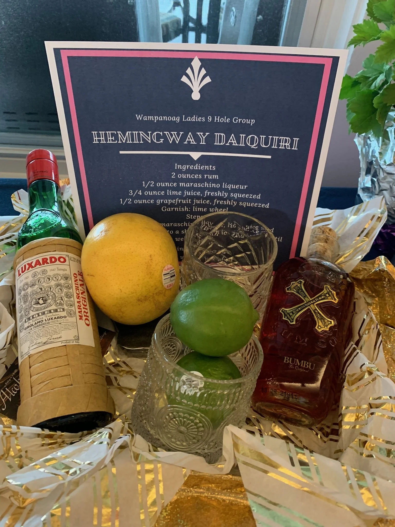 A sign for Wampanoag Ladies 9 Hole Golf Group featuring a recipe for a Hemmingway Daiquiri with ingredients and garnish mentioned. The sign is surrounded by a green lime, yellow lemon, a bundle of grapes, and a bottle of Luxardo Maraschino Liqueur, w