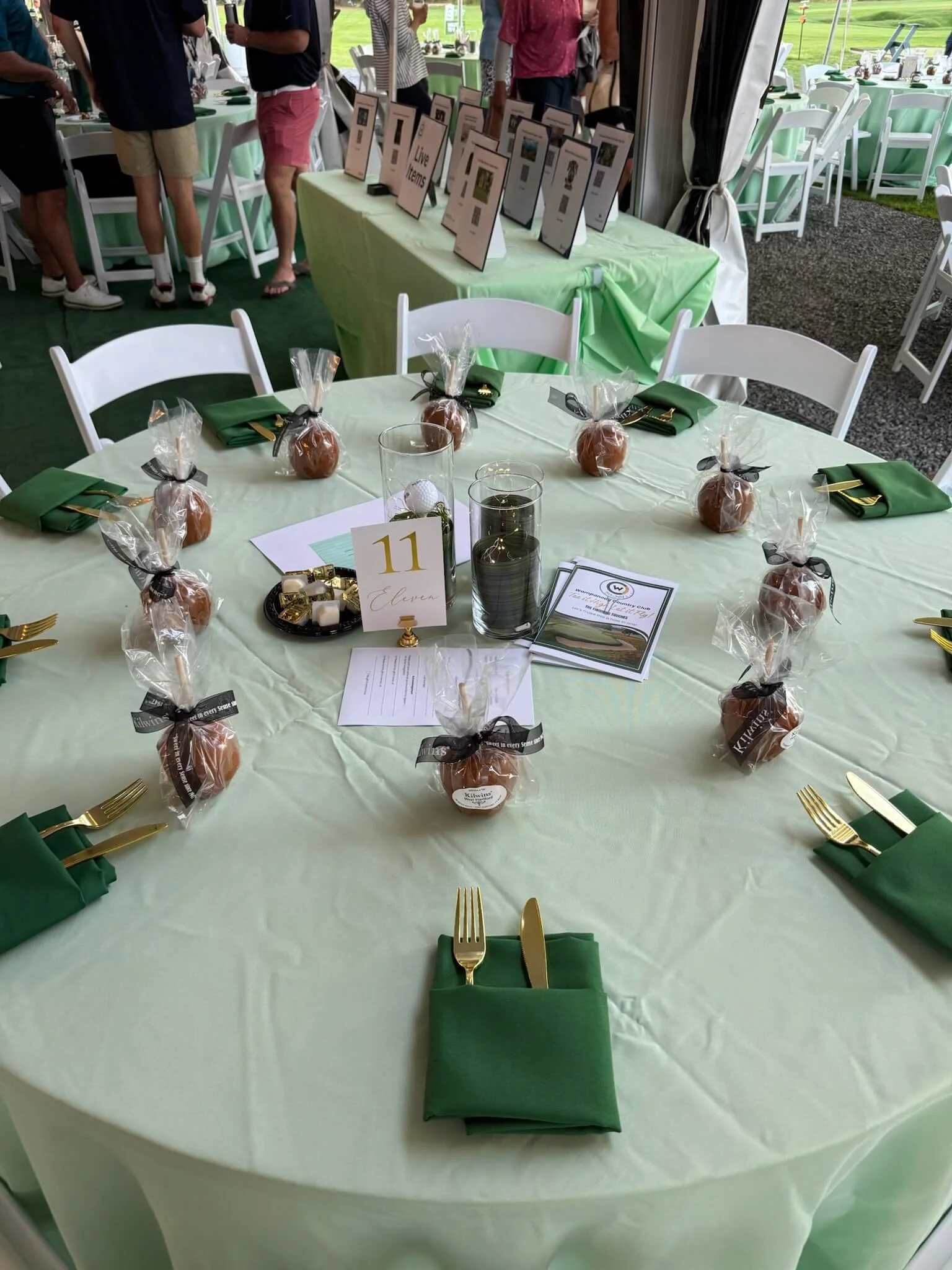 A round banquet table with a light green tablecloth, set with gold utensils, green napkins, and small caramel treats wrapped in clear plastic with black ribbons. In the center, there are two glass candle holders and a table number card with the number 11. Behind the table, there is a display of signs and multiple standing people, with a green outdoor field visible outside the tent.