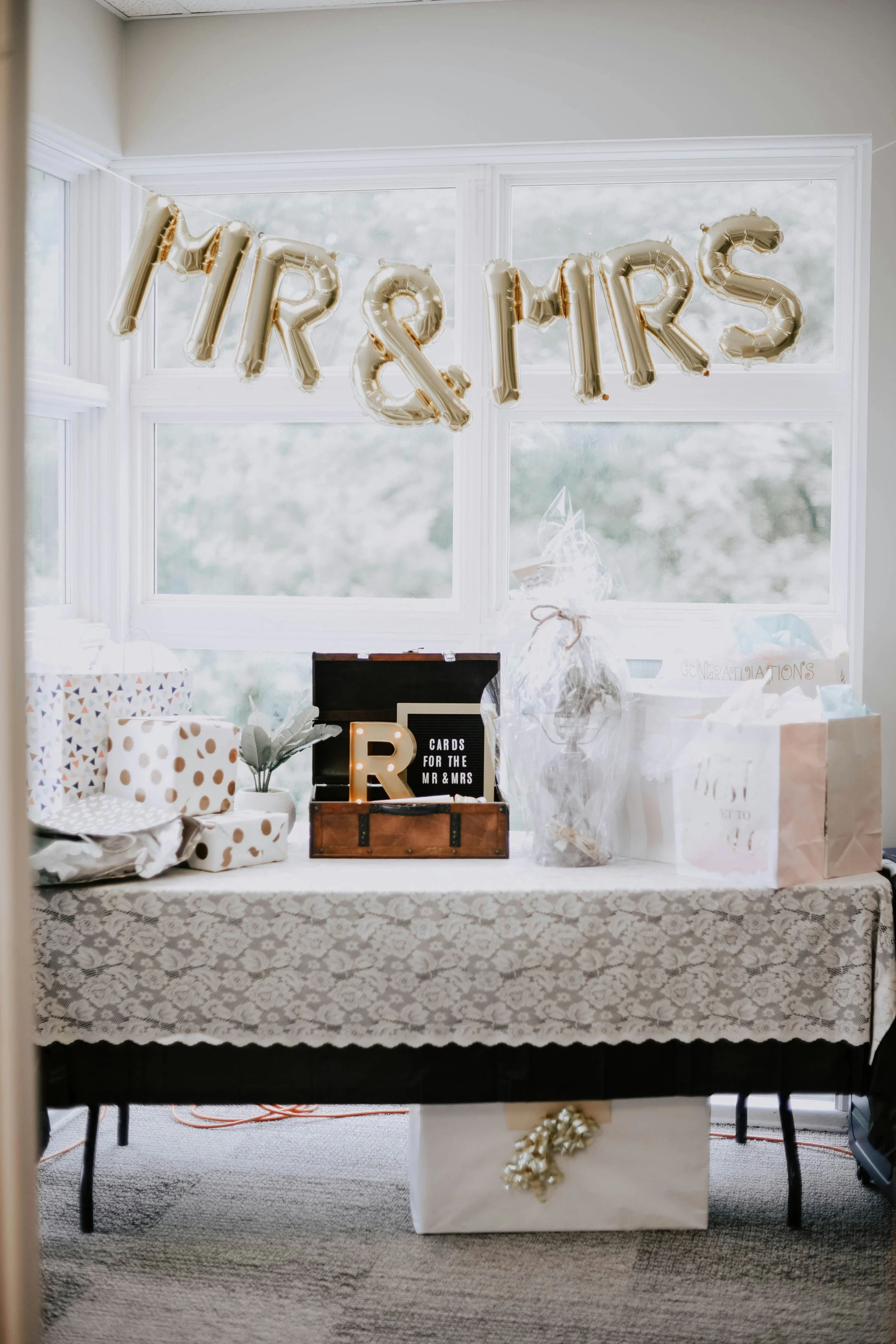 Table decorated for a celebration with gold letter balloons spelling out 'MR & MRS' hanging in front of a large window. The table is covered with a patterned tablecloth and has various wrapped gifts, a small chest with a light-up 'R' and a sign that reads 'CARDS FOR THE MR & MRS'. There is also a white gift bag with gold accents, and decorative items wrapped in transparent paper with ribbons.