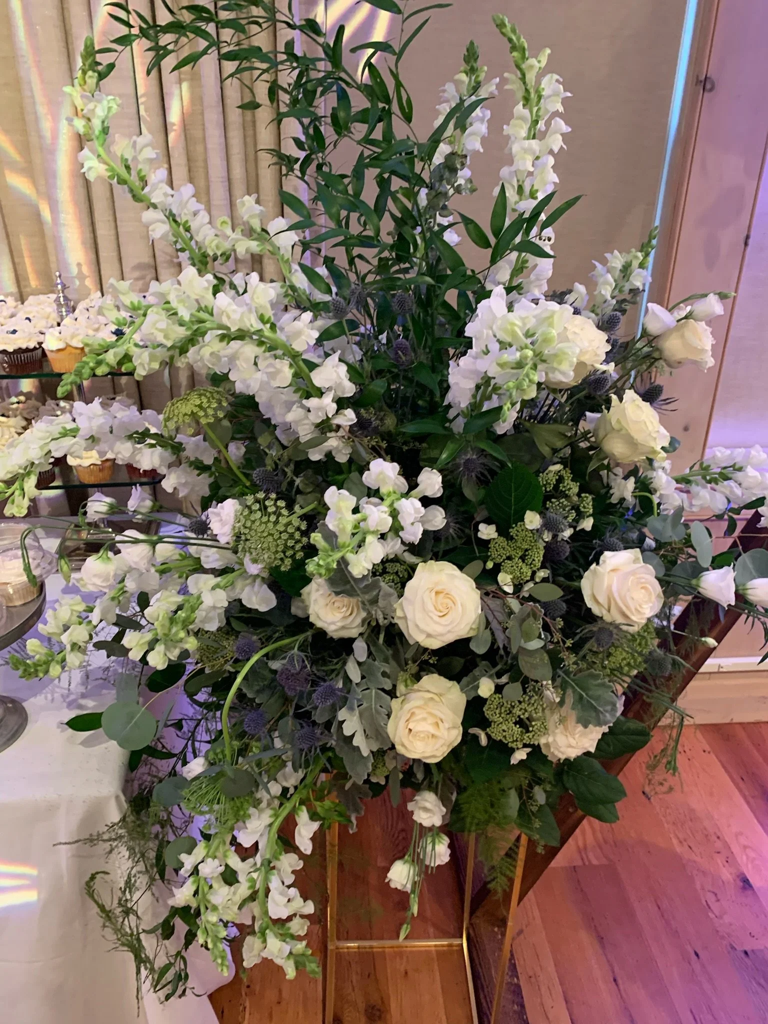 A large floral arrangement featuring white roses, white snapdragons, greenery, and other white flowers, displayed on a wooden table at a formal event.