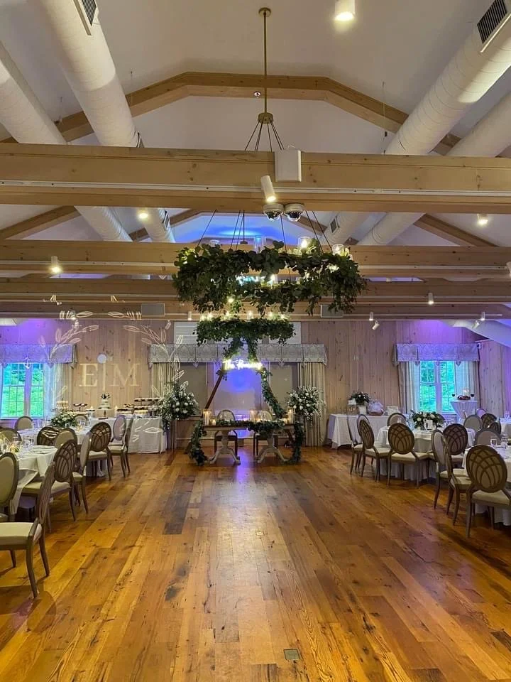 Wedding reception hall decorated with round tables covered in white tablecloths, surrounded by wooden chairs with white cushions. A wooden arch adorned with greenery and flowers is centered at the back of the room. Green floral arrangements are on the tables and at the front. Wooden beams and white air ducts are visible on the ceiling, and windows with curtains are on the sides. The dance floor is in the center with a wooden floor.