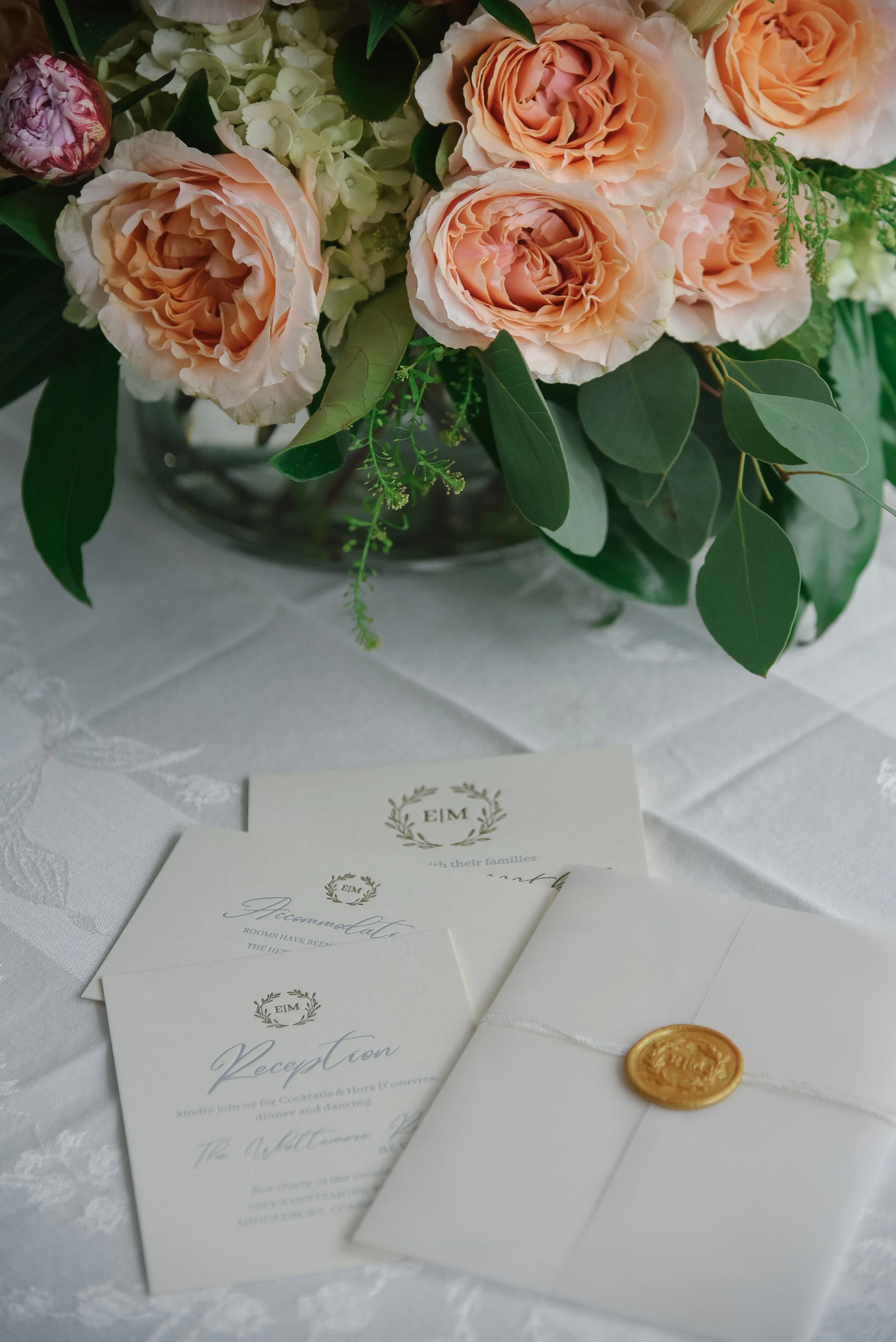 A floral arrangement with pink and white roses, green leaves, and white hydrangeas in a glass vase on a table. In front are wedding reception cards on a white tablecloth. A gold wax seal is visible on the paper.